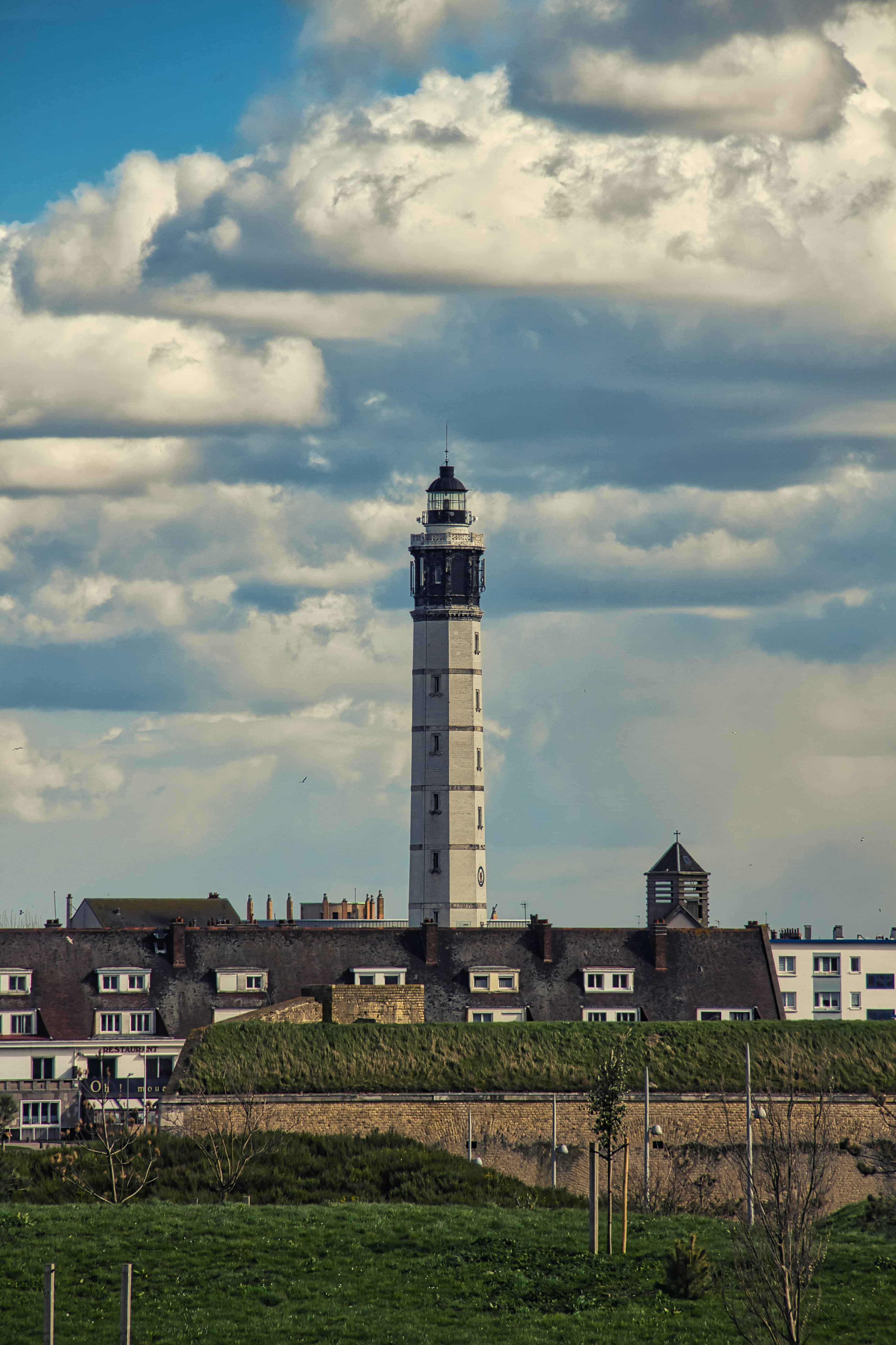 The tall banded lighthouse of Calais rising above the town's rooftops and the grassy ramparts, under a sky of broken cloud. The marathon finishes on the seafront below it.