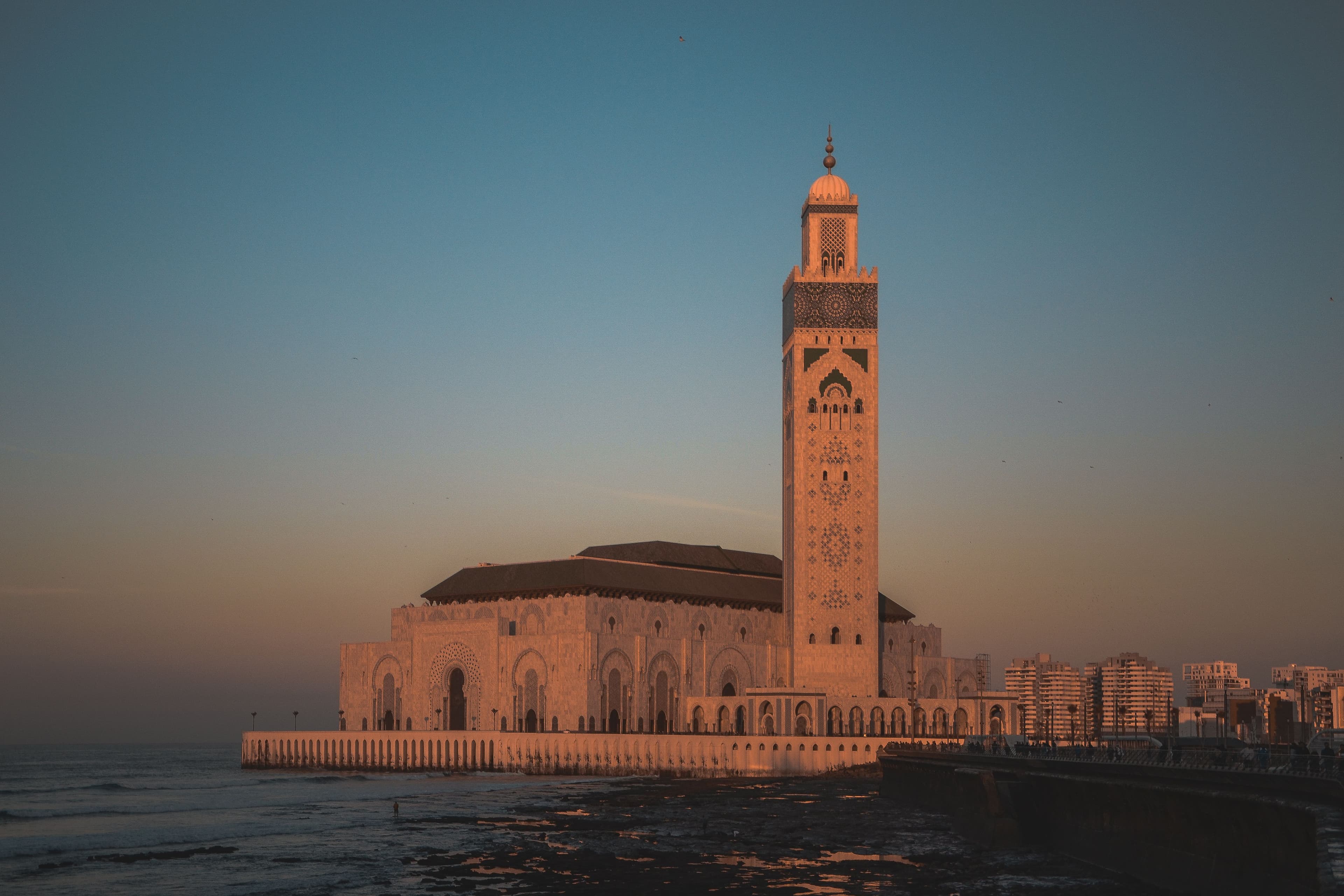 The Hassan II Mosque at sunset, its minaret rising above the Atlantic on the Casablanca waterfront, the sky fading from blue to warm amber