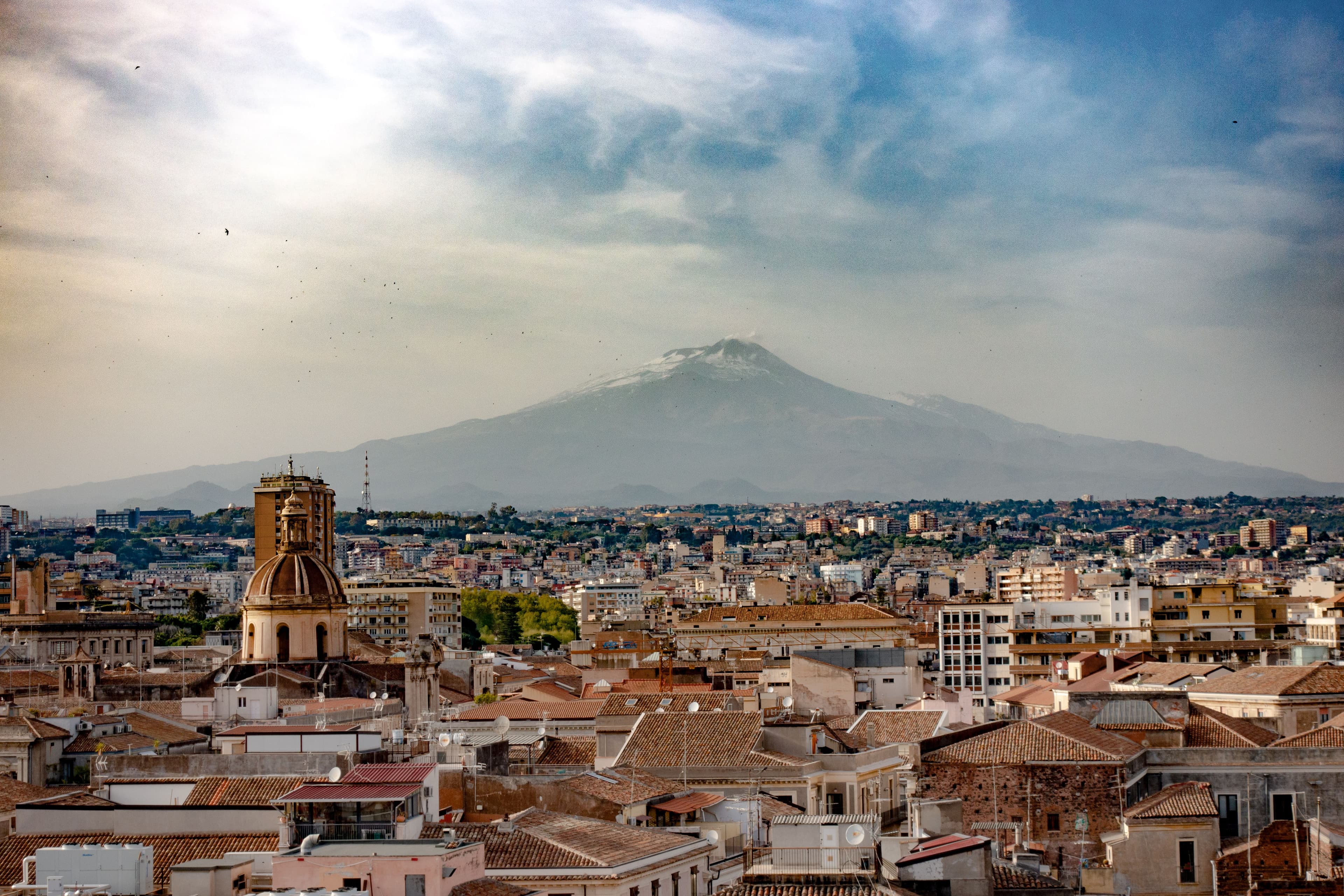 Terracotta rooftops and a domed church in central Catania under a hazy sky, with snow on the summit of Mount Etna in the distance