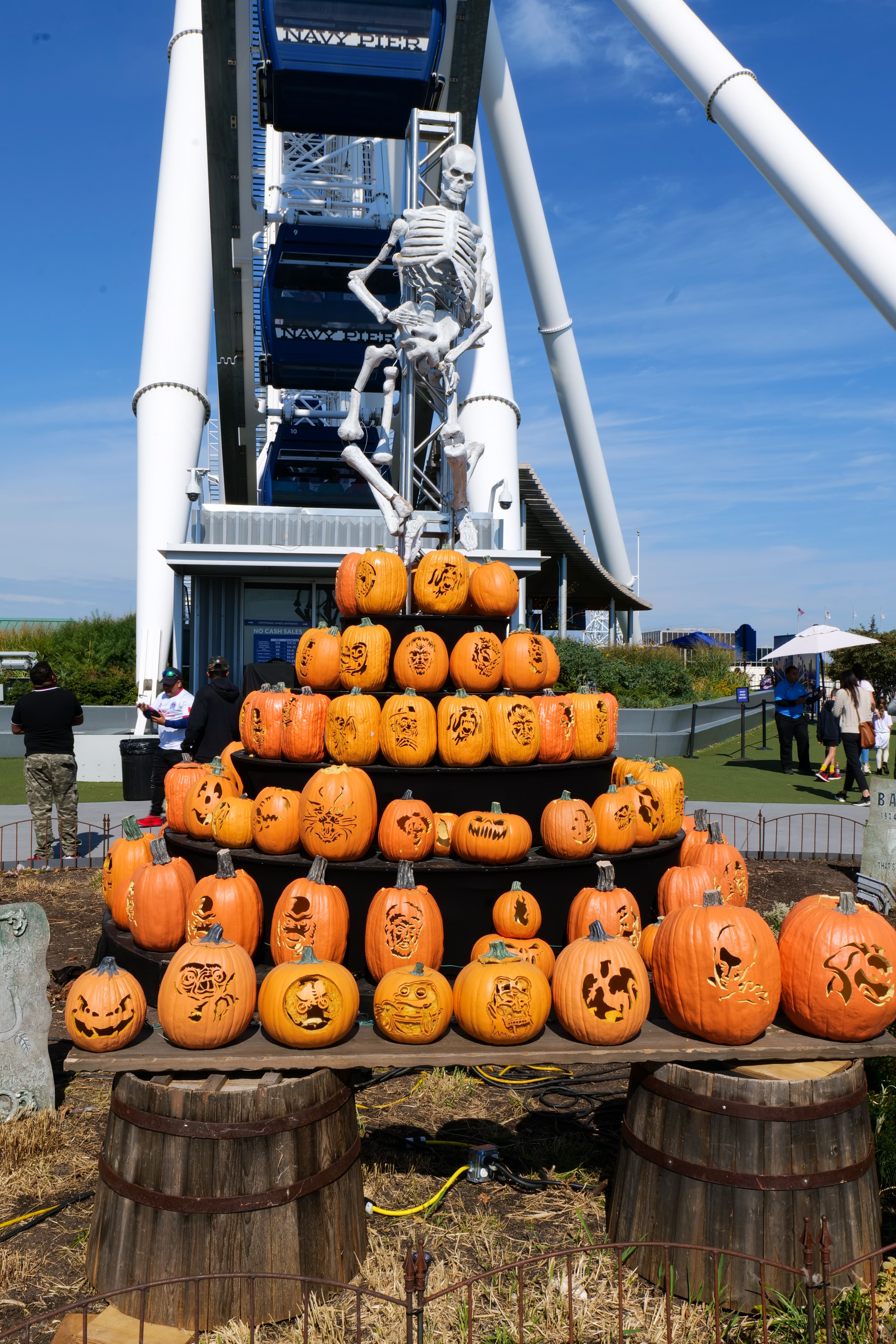A stepped pyramid display of carved and decorated jack-o'-lanterns at Navy Pier, Chicago, in mid-October, with a large skeleton figure at the top and the Navy Pier Ferris wheel visible behind against a clear blue sky