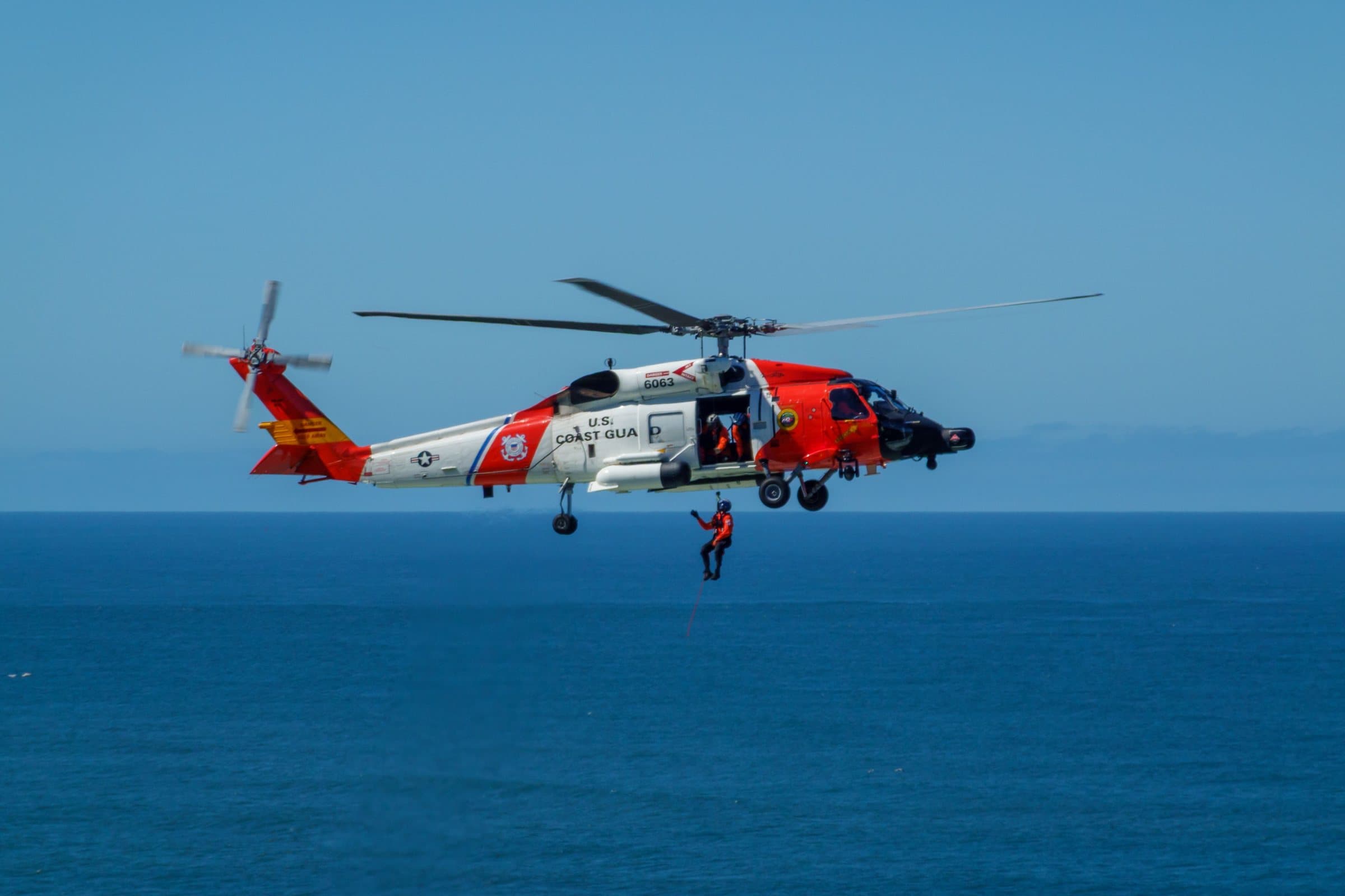 An orange and white Coast Guard helicopter hovering over open sea under a clear blue sky while a rescue swimmer is lowered on a line