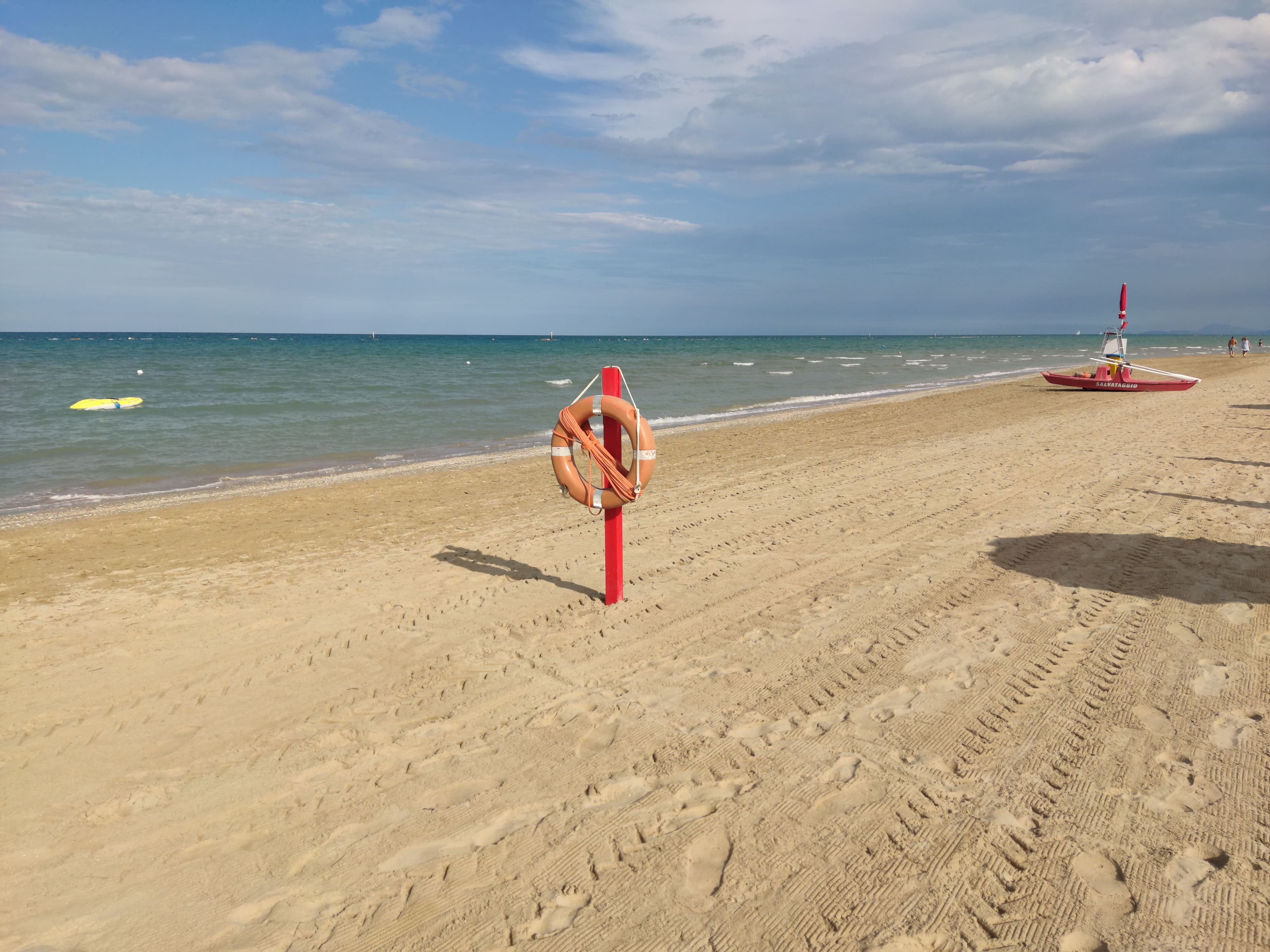 A deserted sandy beach at Fano on the Adriatic with a red lifebuoy on a post, a lifeguard rescue boat and calm turquoise sea under a broken sky