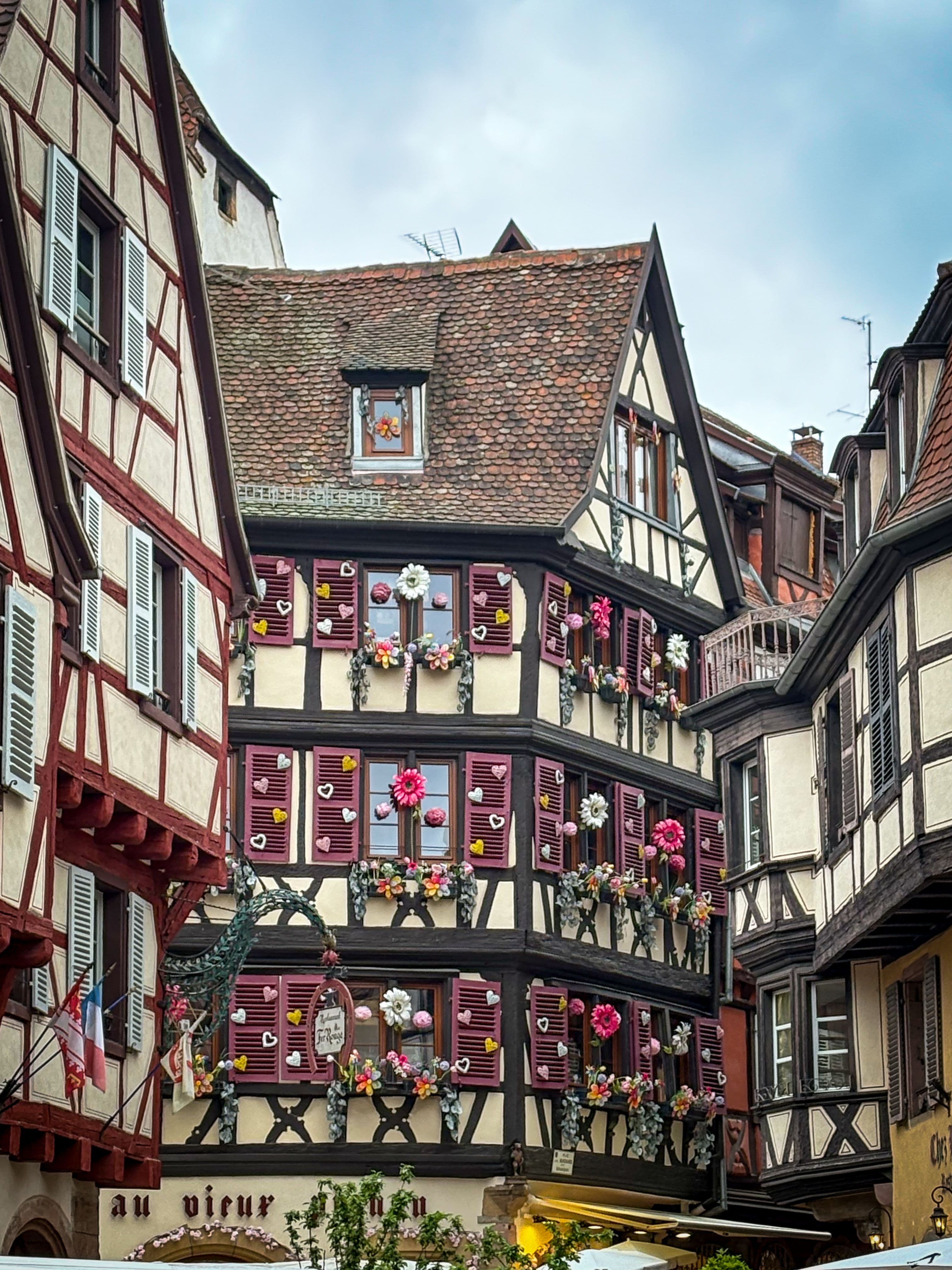 A tall half-timbered house in Colmar's old town with burgundy shutters covered in knitted flowers and hearts, a restaurant sign below and the French and Alsatian flags on the building opposite.
