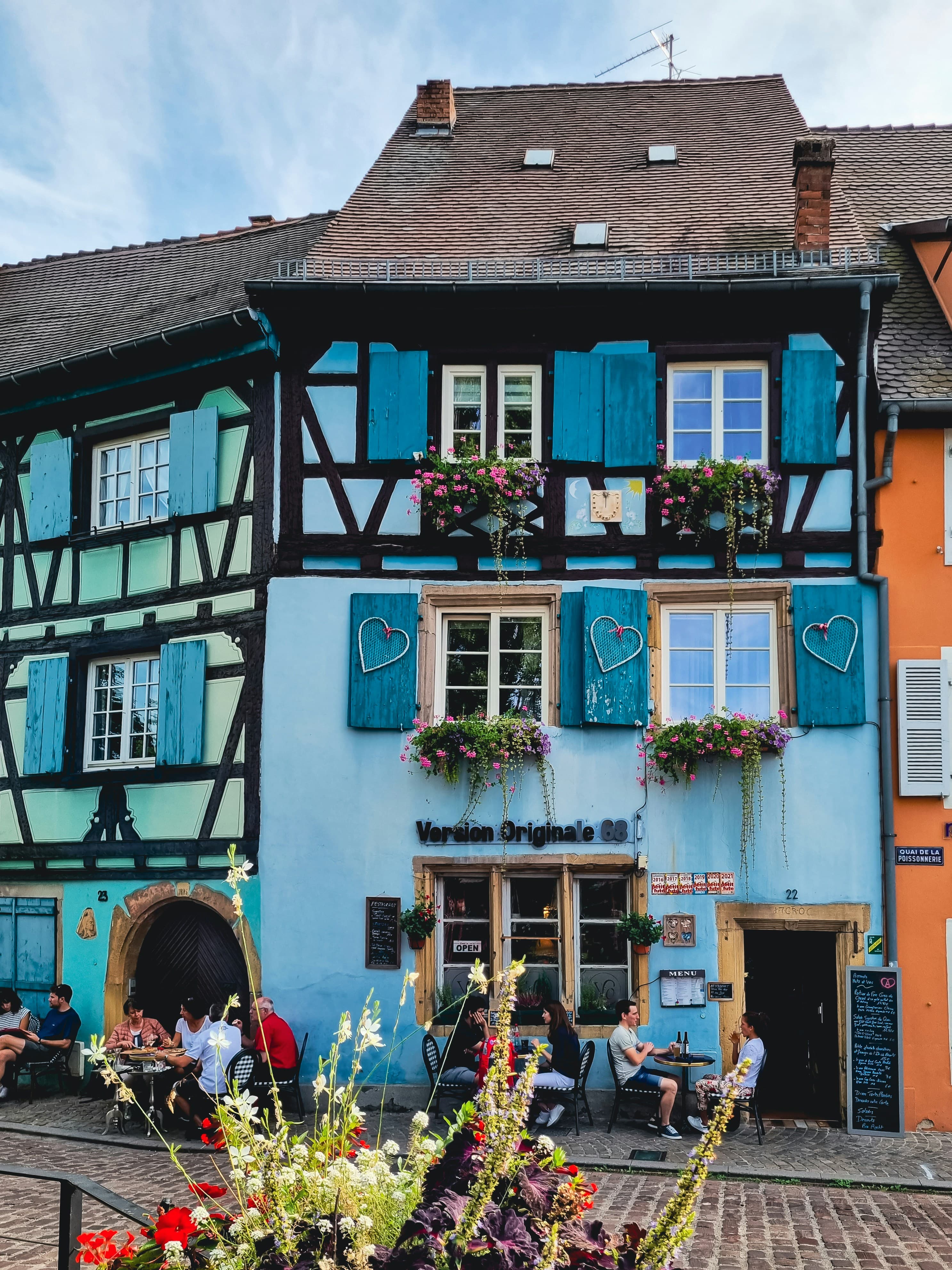 A pale blue half-timbered building on Quai de la Poissonnerie in Colmar, its shutters hung with woven hearts and window boxes of geraniums, with diners at pavement tables on the cobbles below.
