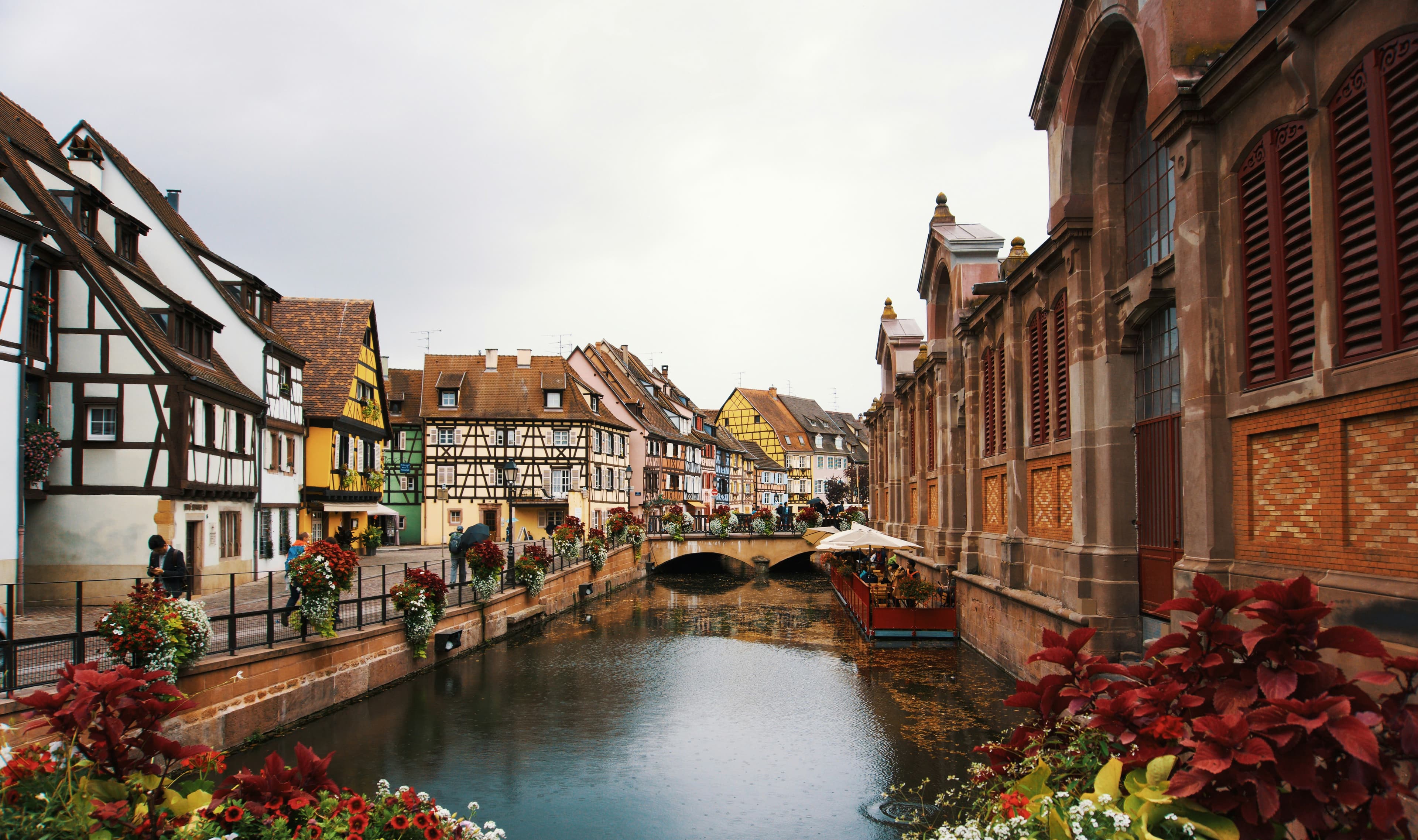 The canal at Petite Venise in Colmar, lined with half-timbered houses in yellow, pink and green, flower boxes along the railings and the sandstone covered market on the right. The marathon starts and finishes a short walk away at Place Rapp.