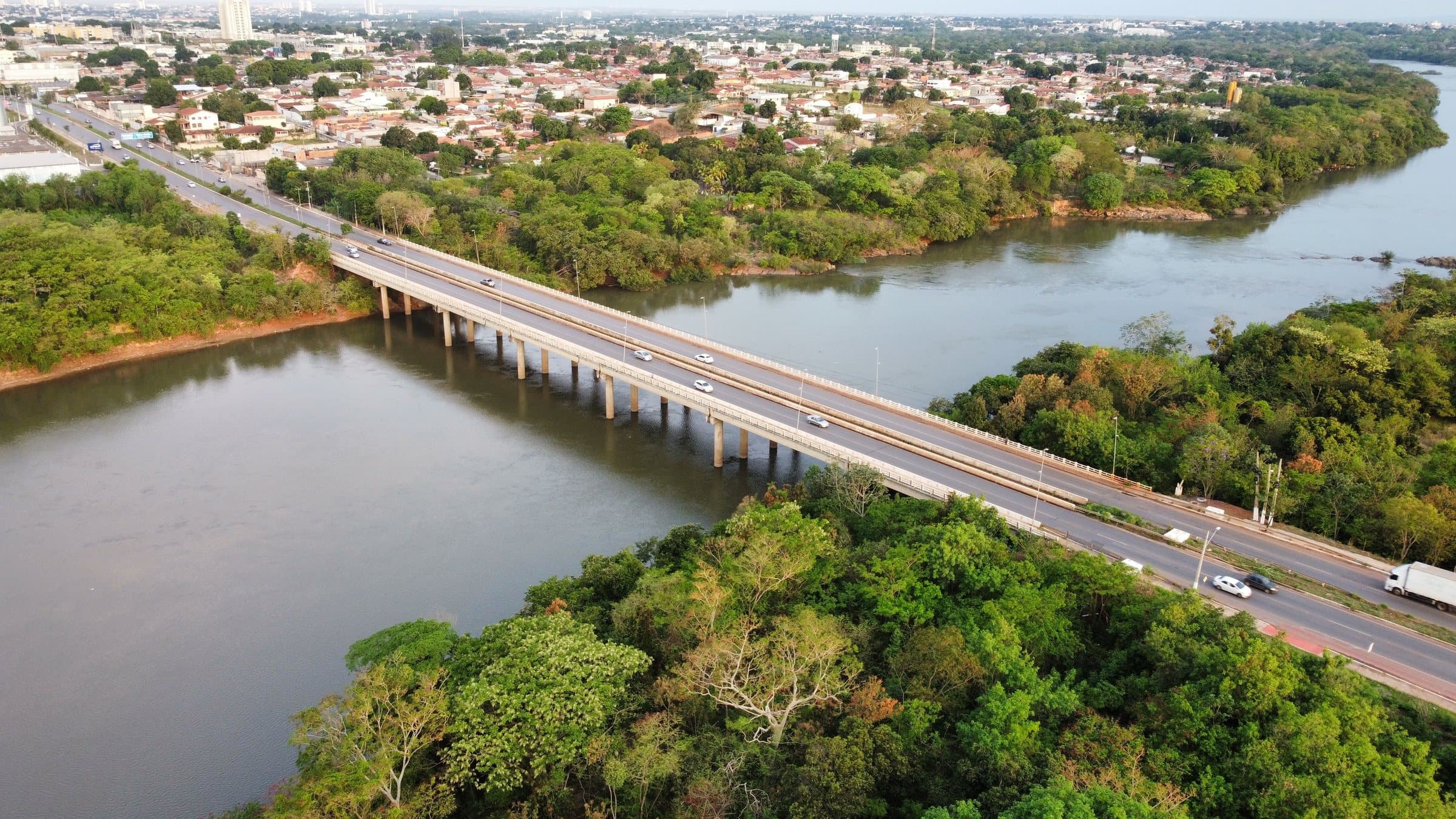Aerial view of a road bridge crossing a wide river in Cuiabá, with green riverbank trees and the city beyond in warm light