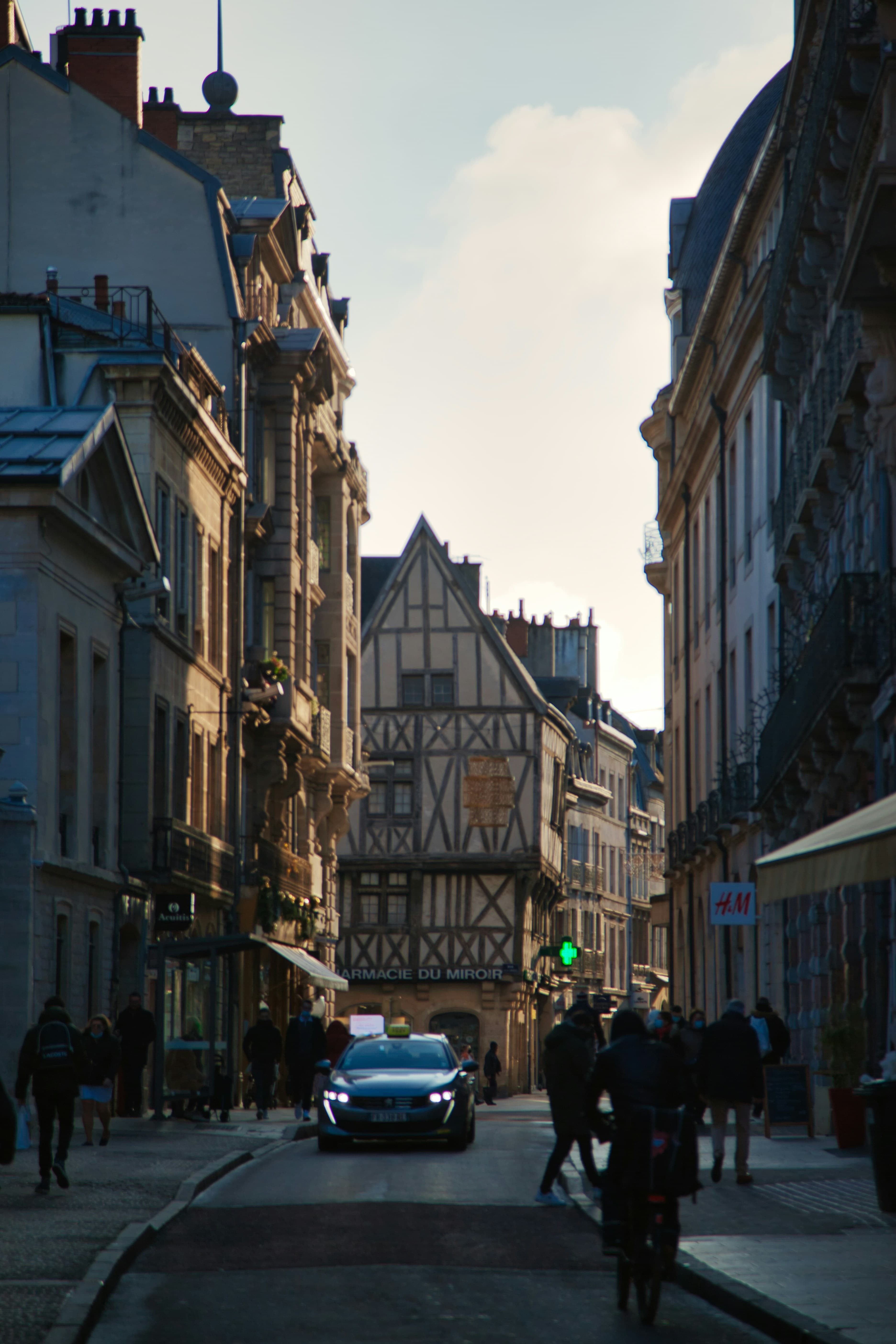 A narrow Dijon shopping street in low winter sun, with a timber-framed medieval building housing the Pharmacie du Miroir at the far end and a taxi waiting in the road.