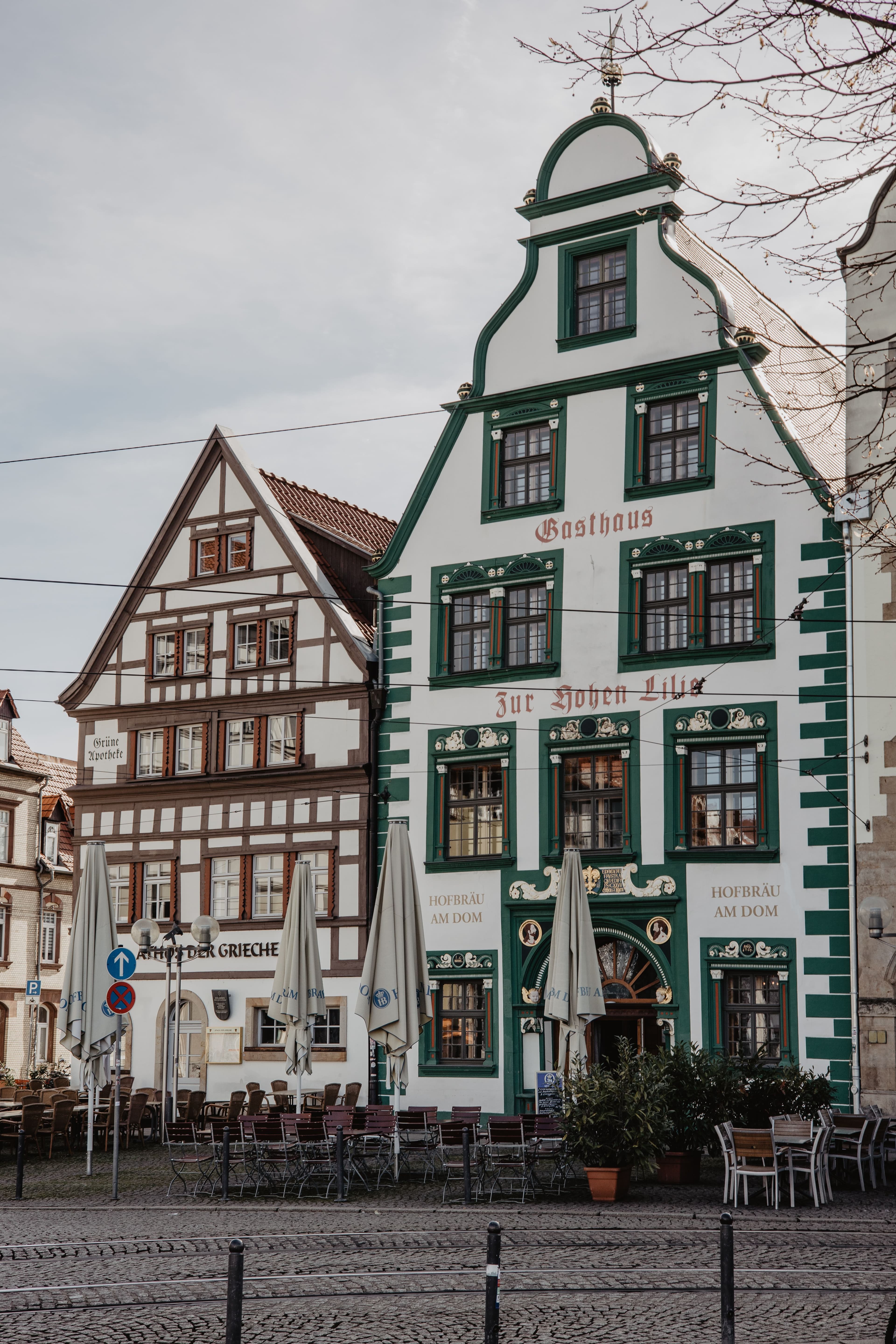 The green and white gabled facade of the Gasthaus Zur Hohen Lilie on Erfurt's Domplatz, beside a half-timbered building housing the Grüne Apotheke, with cafe tables set out on the cobbles
