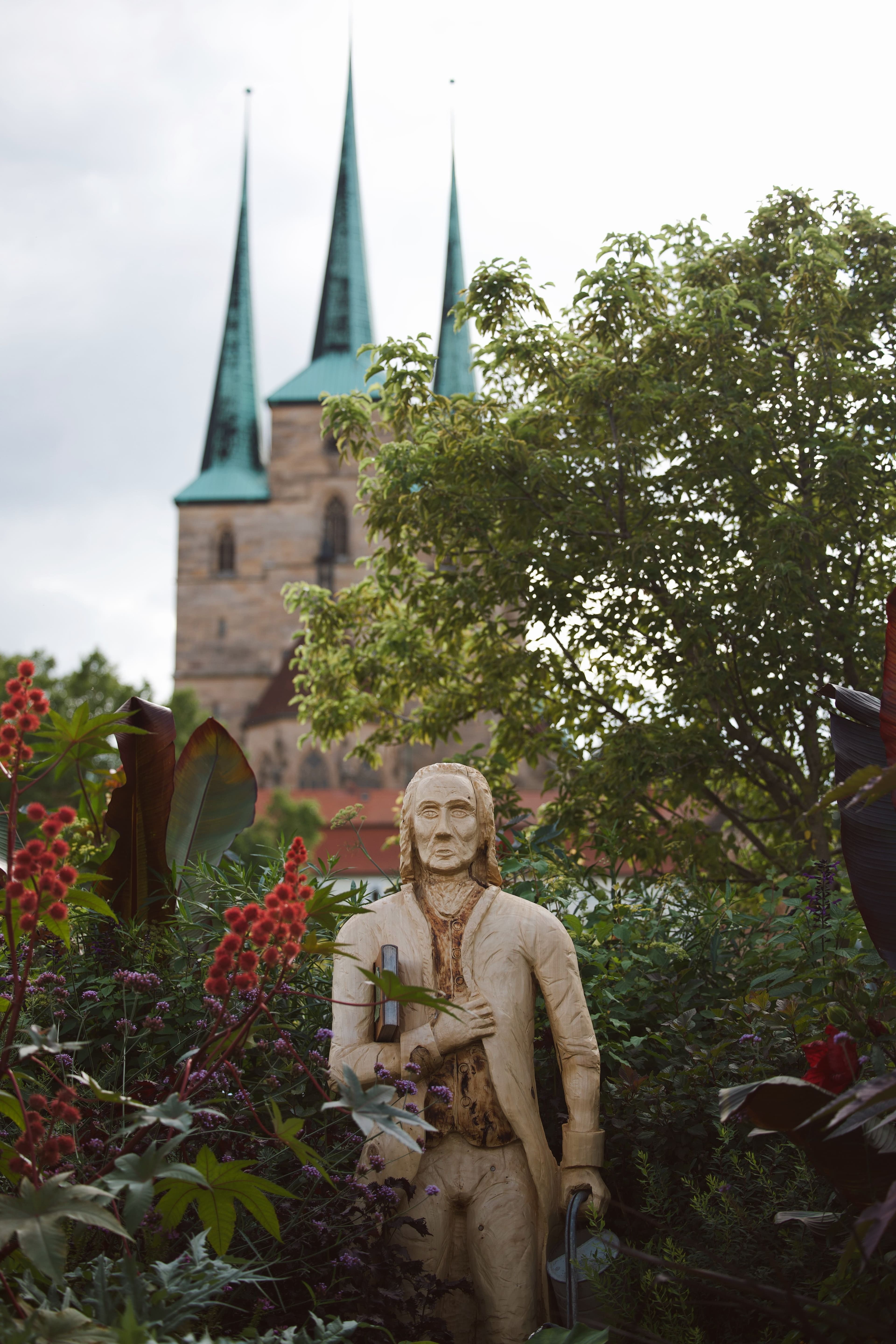 The three green spires of the Severikirche rising above a late-summer garden in Erfurt, with a carved wooden figure holding a book in the foreground