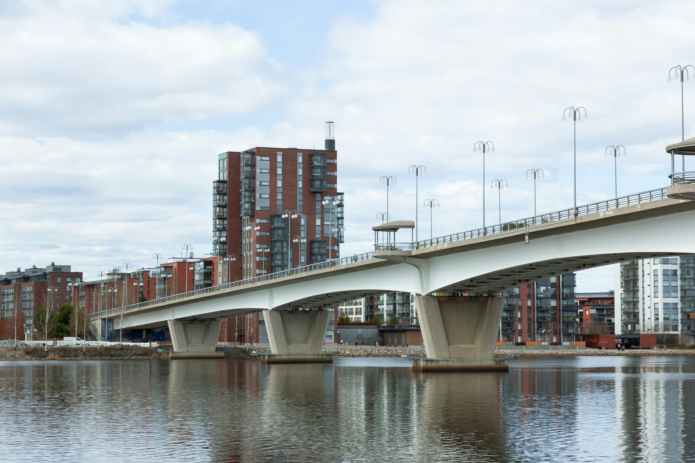A white concrete road bridge on three piers crossing a lake in Jyväskylä, with red-brick and glass apartment towers on the shore under a cloudy sky