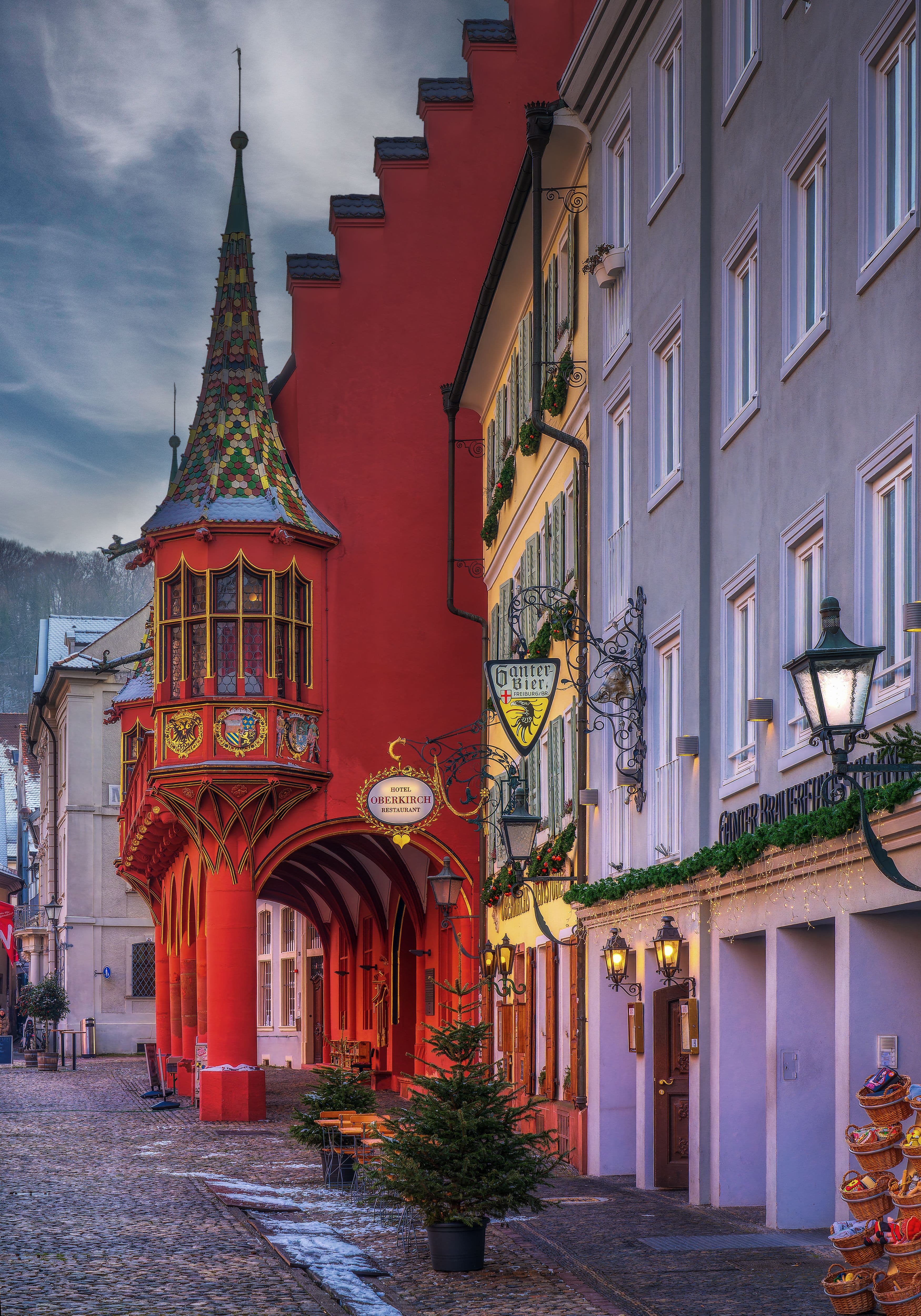 The red arcaded Historisches Kaufhaus on Münsterplatz in Freiburg, its tiled oriel turret lit at dusk. Münsterplatz sits at the heart of the old-town section the marathon passes twice.