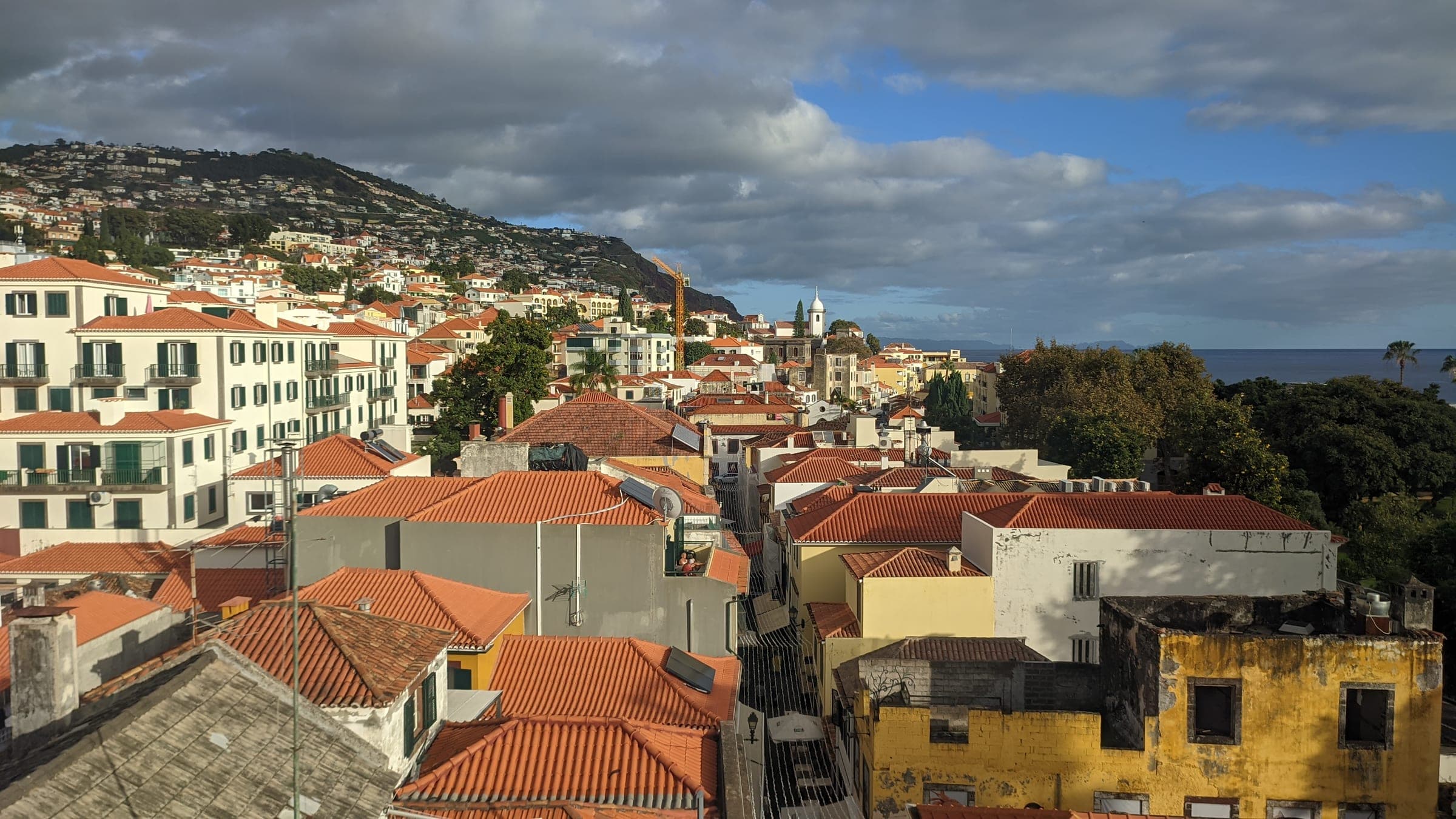 Terracotta rooftops of central Funchal under a broken sky, with white houses climbing the hillside above the city and the Atlantic on the right, the city the Funchal Marathon finishes in