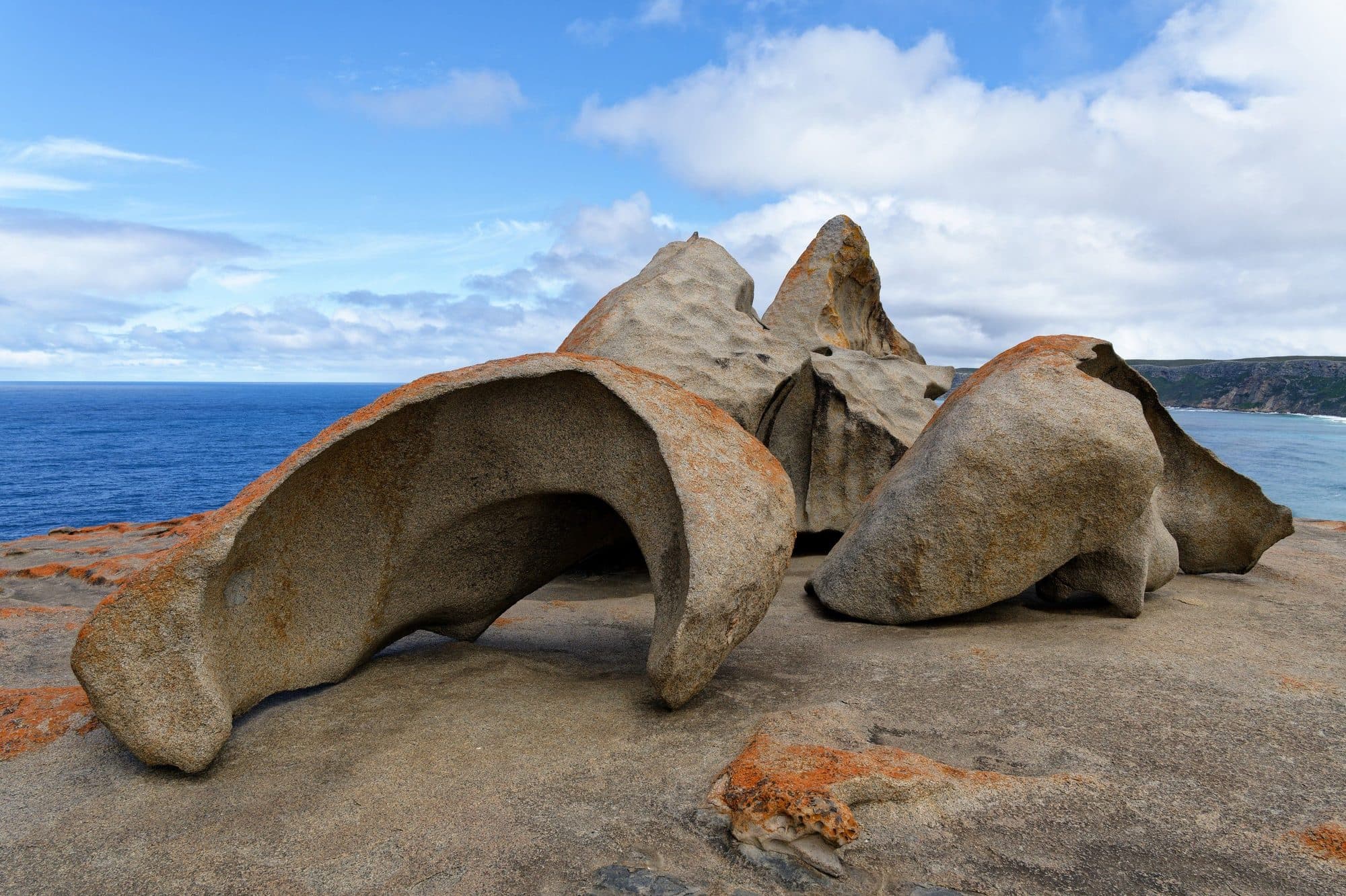 Wind-sculpted granite boulders with orange lichen at Remarkable Rocks in Flinders Chase National Park, with the Southern Ocean and a headland behind