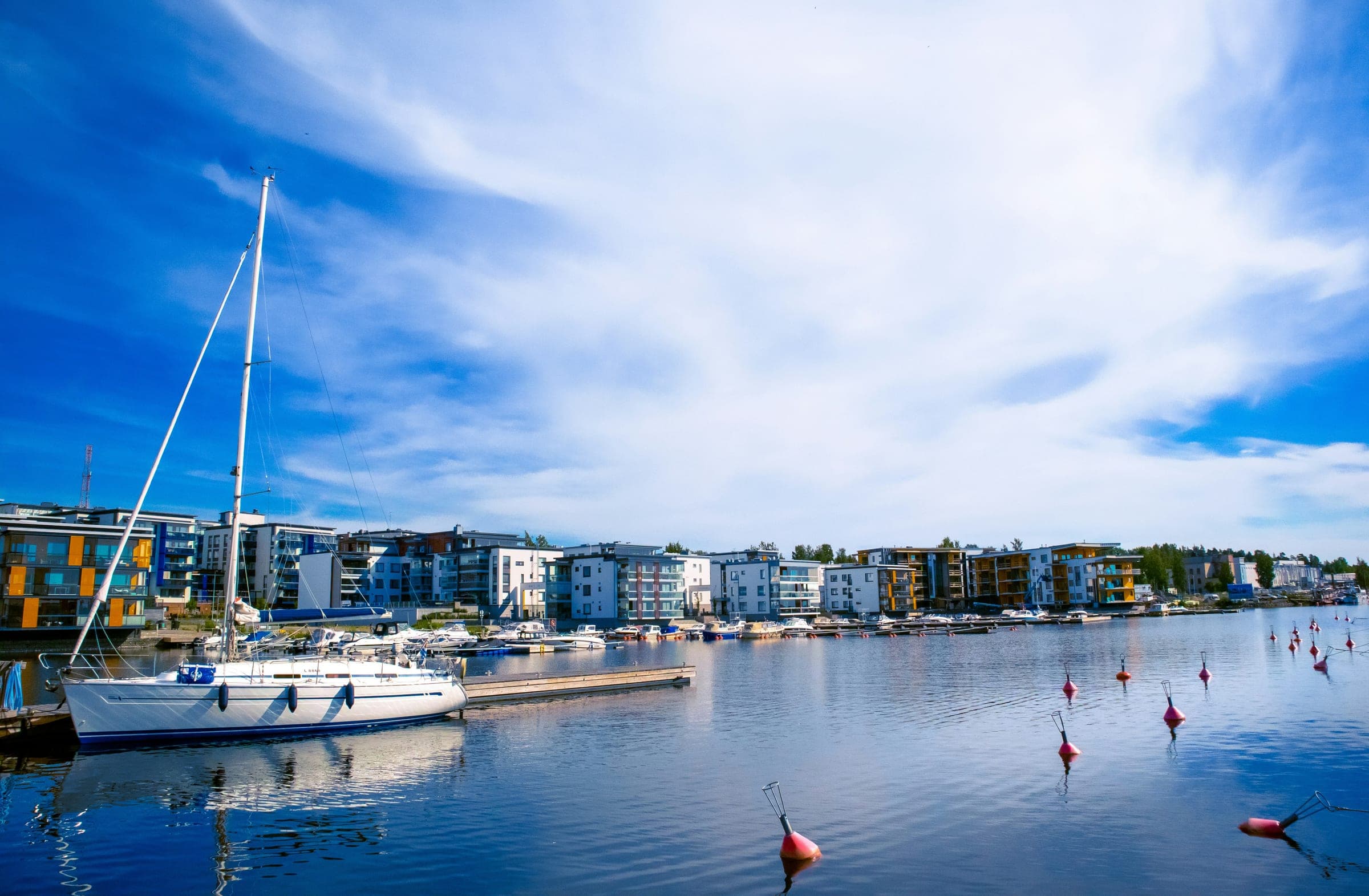A white sailing yacht moored at a wooden jetty in a lakeside marina in Kuopio, with modern apartment blocks along the shore and red buoys on the water under a bright blue sky