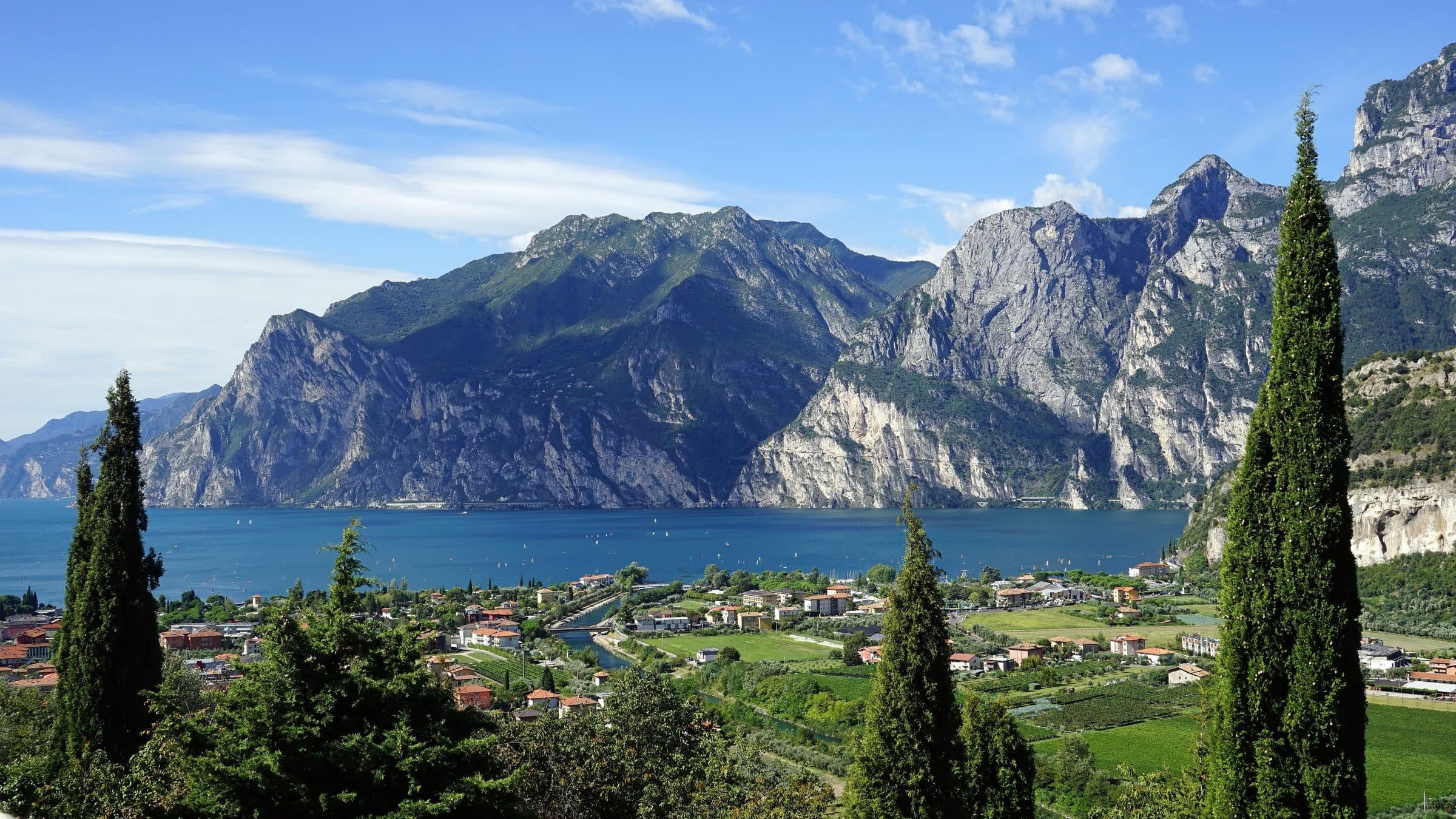 The northern end of Lake Garda, with the limestone cliffs of the western shore across the water, cypress trees in the foreground and a lakeside town below