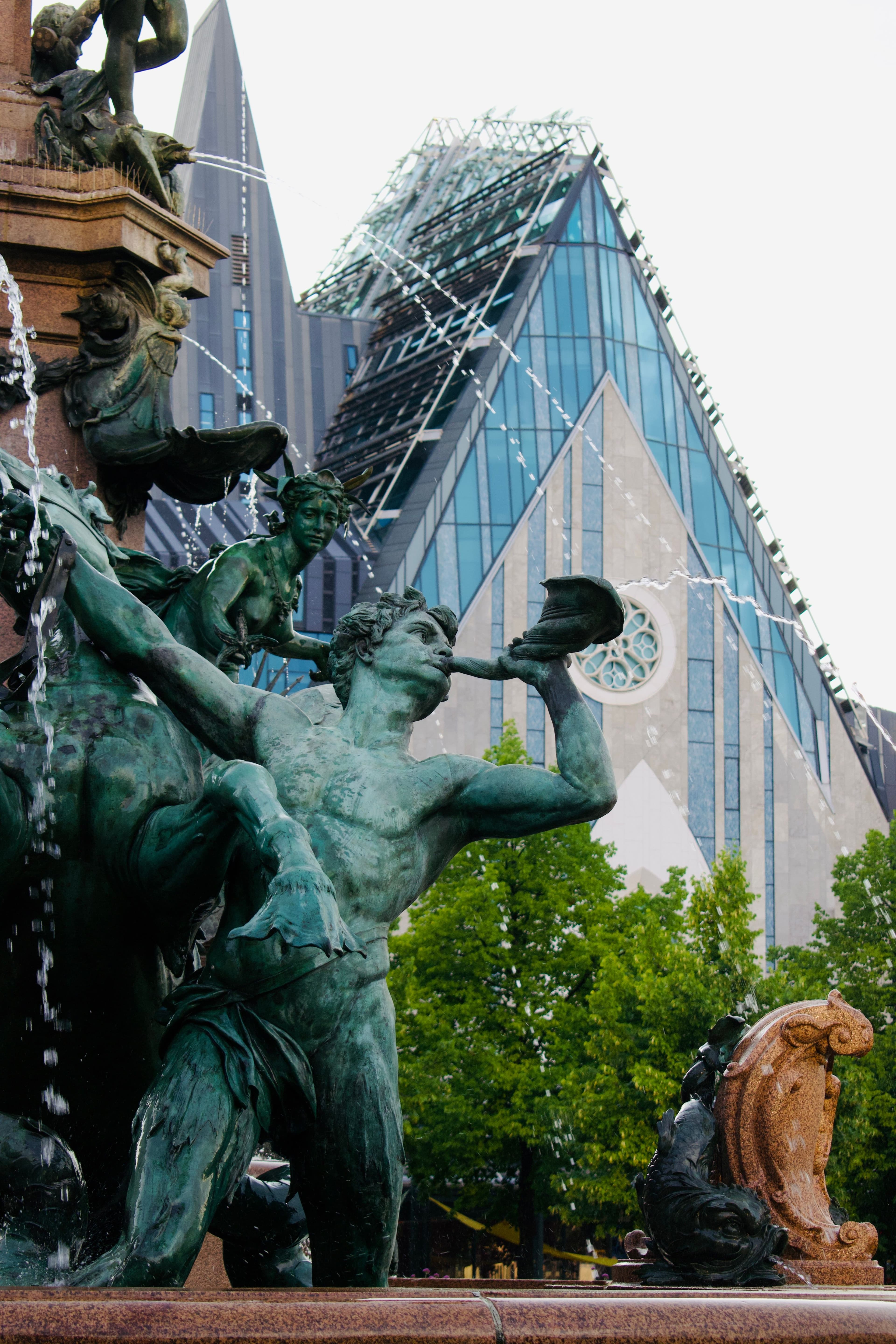 The bronze figures of the Mendebrunnen fountain on Augustusplatz in Leipzig, with the glass and stone Paulinum of the university behind. The marathon reaches Augustusplatz at about km 2.3 on each lap.