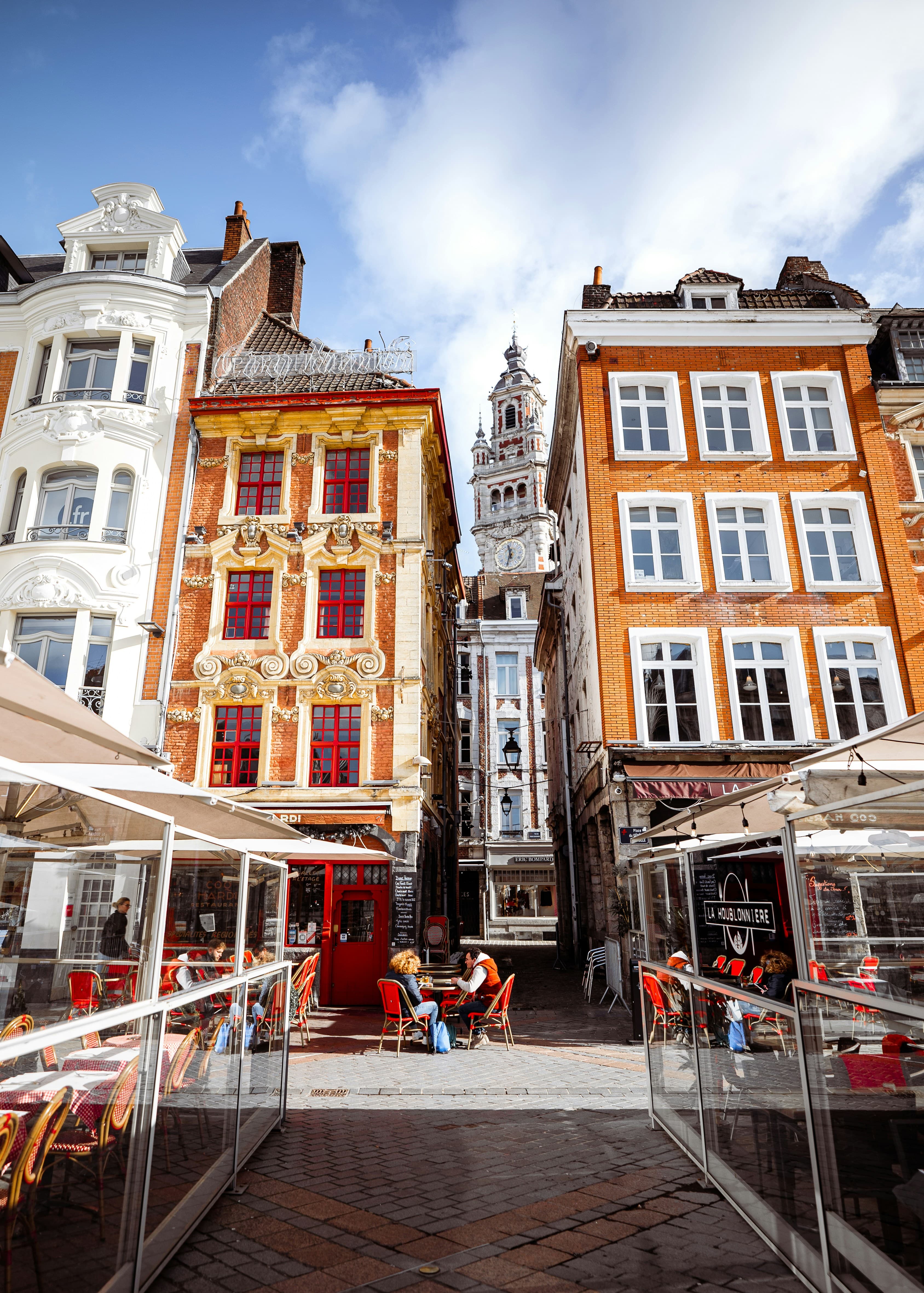 Brick and stone Flemish townhouses at a corner of Lille's Grand'Place, one with ornate red window frames, and between them a narrow lane framing the belfry of the Chamber of Commerce. Café terraces fill the square in the foreground.
