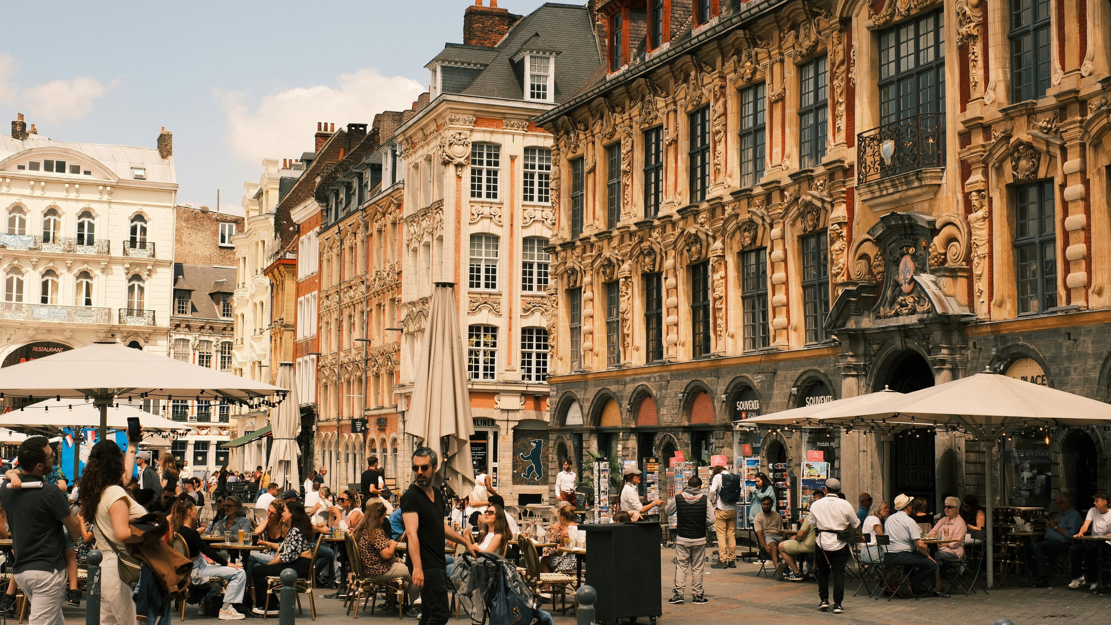 Café terraces on Lille's Grand'Place in front of the carved brick and stone facade of the Vieille Bourse, with Flemish townhouses behind. The 2026 marathon course passes the square at 2.7 km and again at 38.3 km.