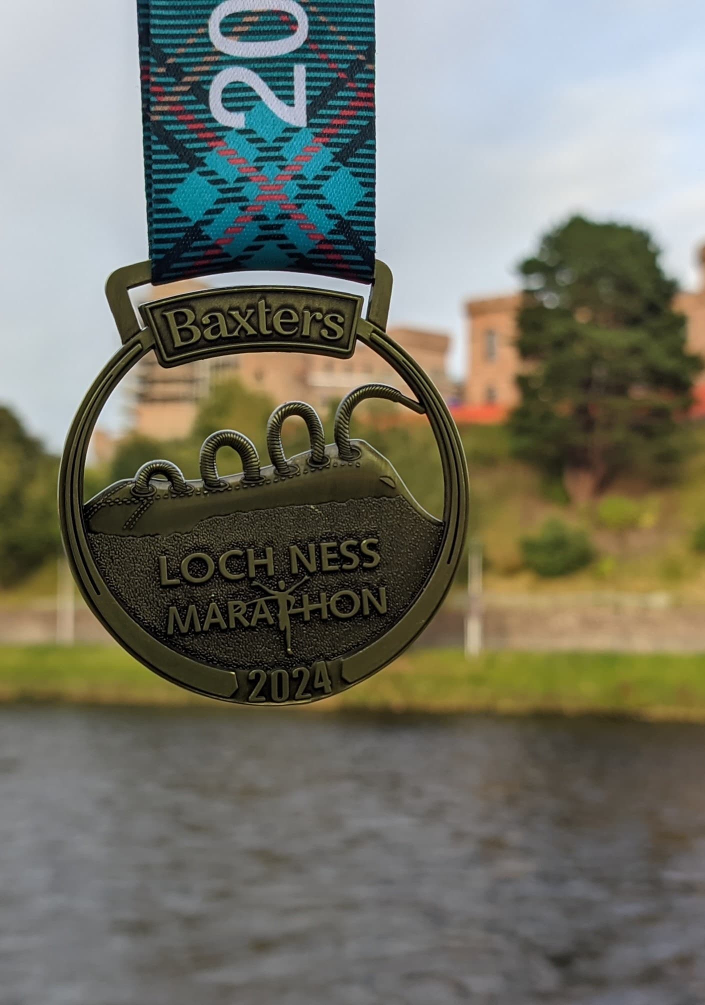 The 2024 Baxters Loch Ness Marathon finisher's medal, shaped like Nessie with laces along its back, photographed beside the River Ness in Inverness