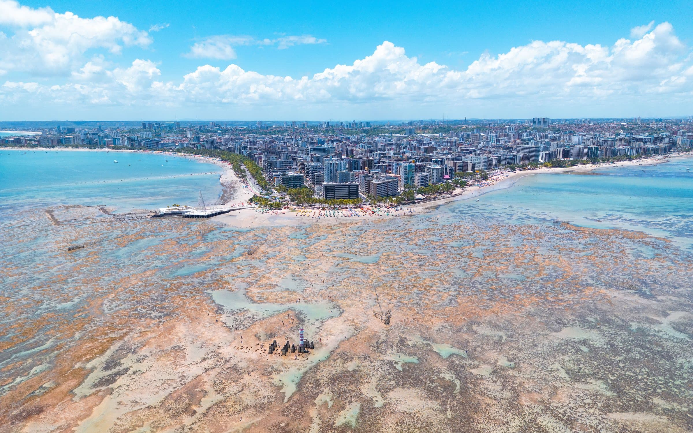 Aerial view of the Pajuçara reef pools at low tide, with the Maceió beachfront and city skyline behind under a blue sky with white clouds