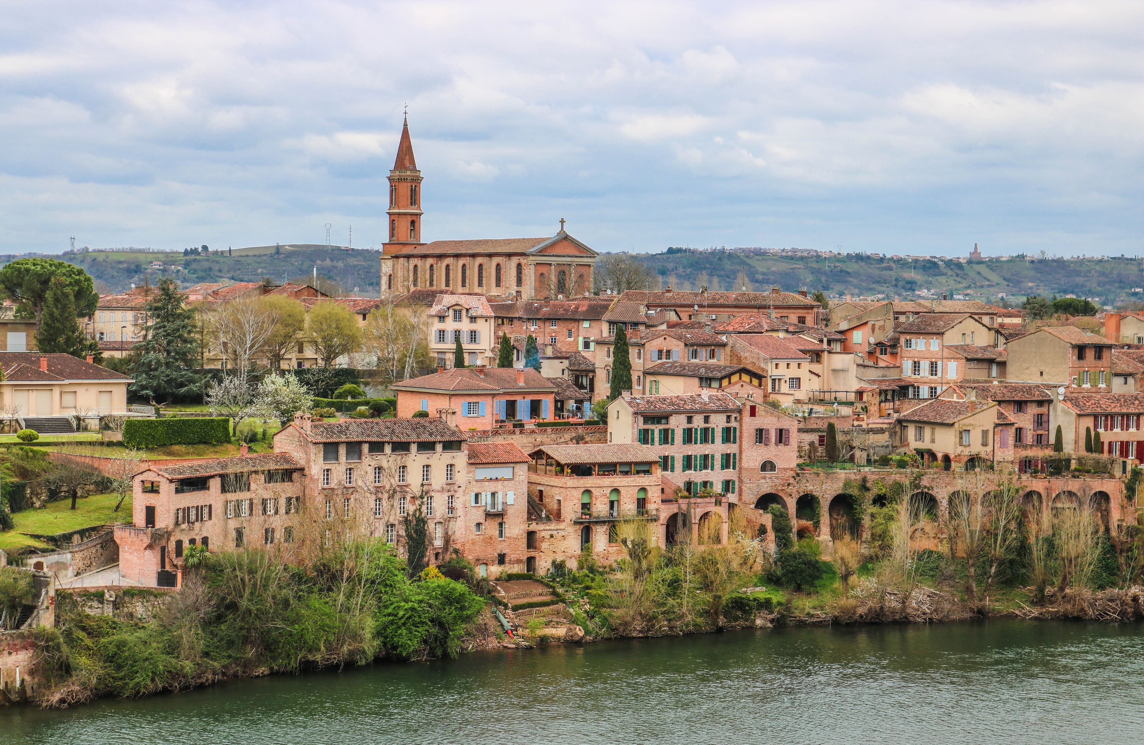 The Madeleine quarter of Albi seen across the Tarn: brick houses with coloured shutters and arcades above the riverbank, and the bell tower of the Église de la Madeleine on the skyline.