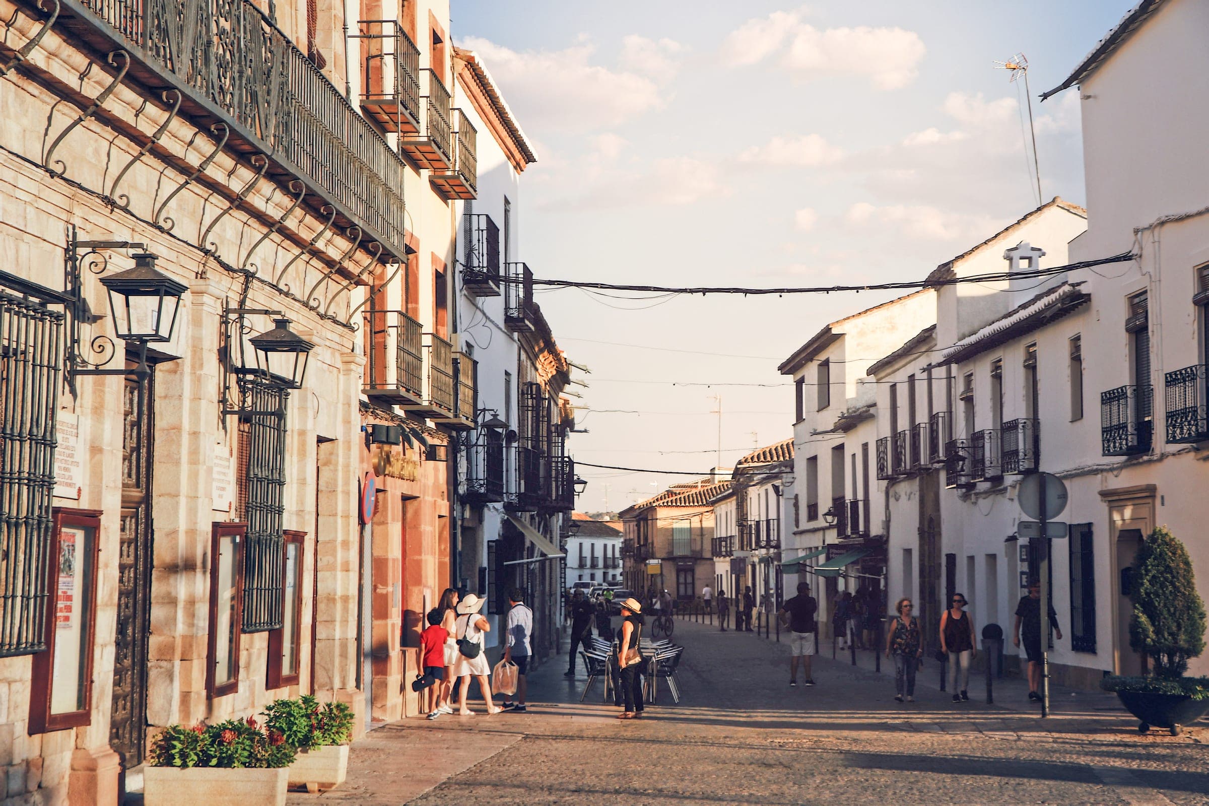 A street in the historic centre of Almagro with whitewashed and stone houses, iron balconies and lanterns, and people walking in low evening sun, near the Plaza Mayor 750 m from the marathon start