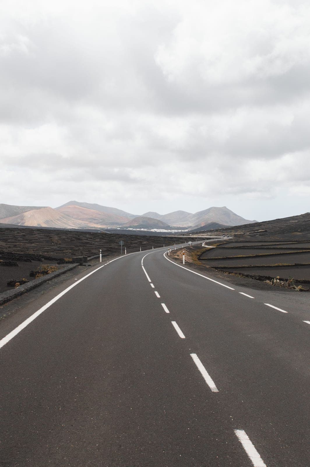 An empty road running through black lava fields towards volcanic cones under a cloudy sky, typical of the Lanzarote interior visited on the Timanfaya day trip