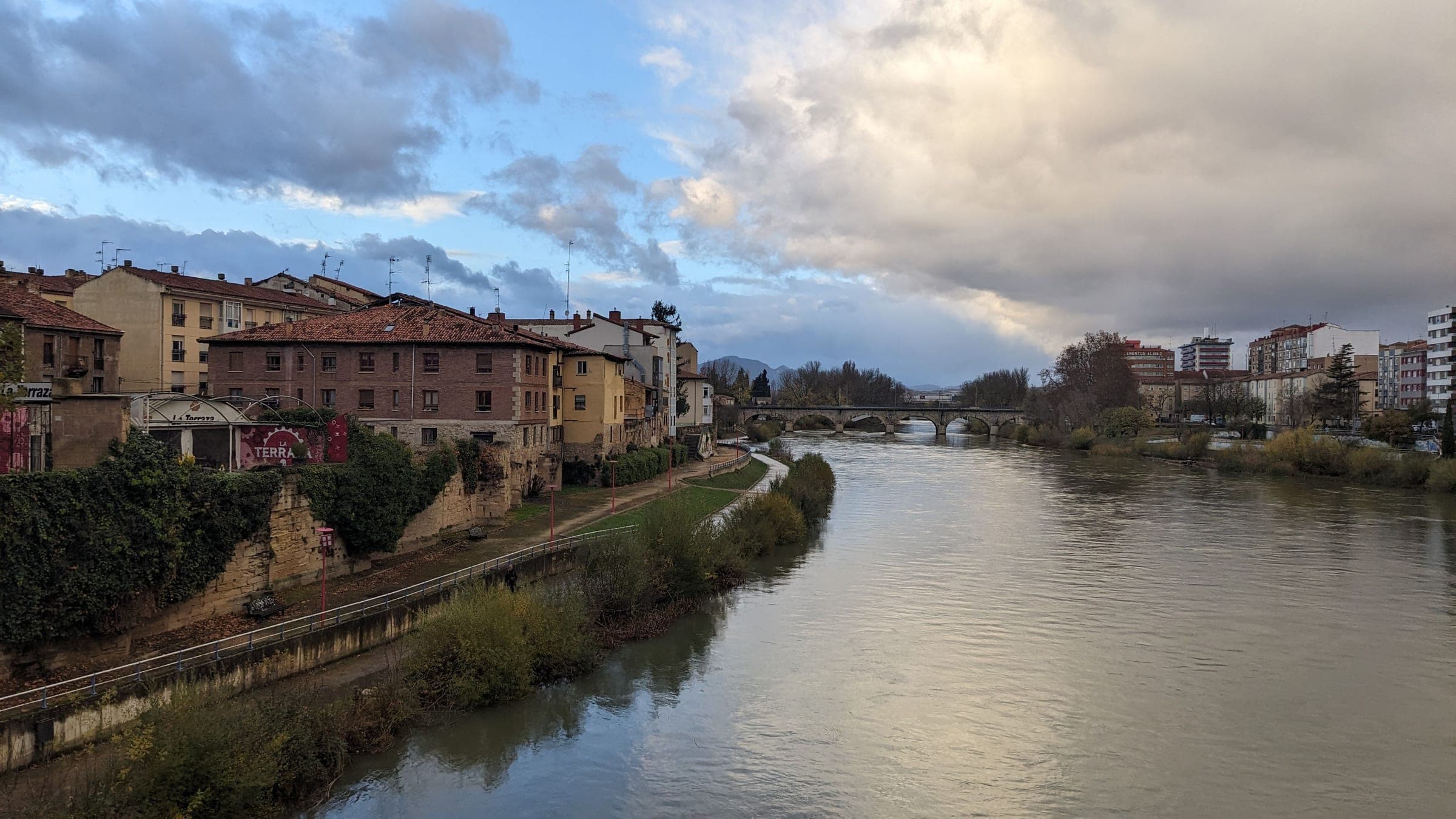 The Ebro at Logroño under a stormy autumn sky, with the old town’s houses and riverside path on the south bank and a stone bridge downstream, the river the marathon crosses on each lap