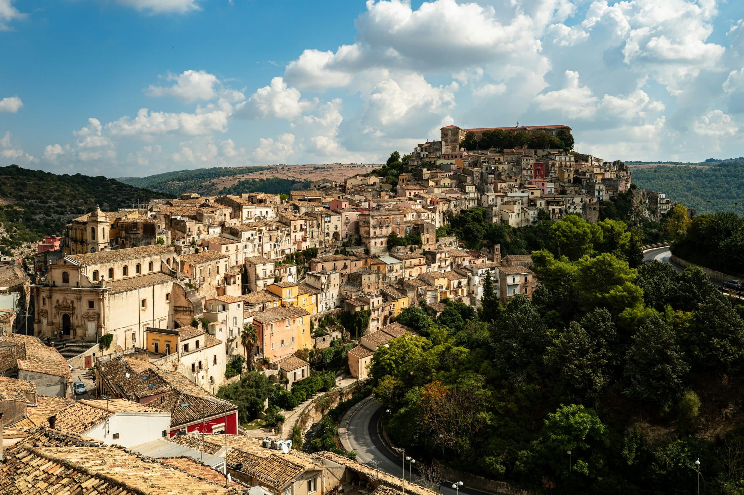 The Baroque hillside town of Ragusa Ibla, stacked up a slope of honey-coloured houses and churches, with green hills behind and a white-clouded blue sky