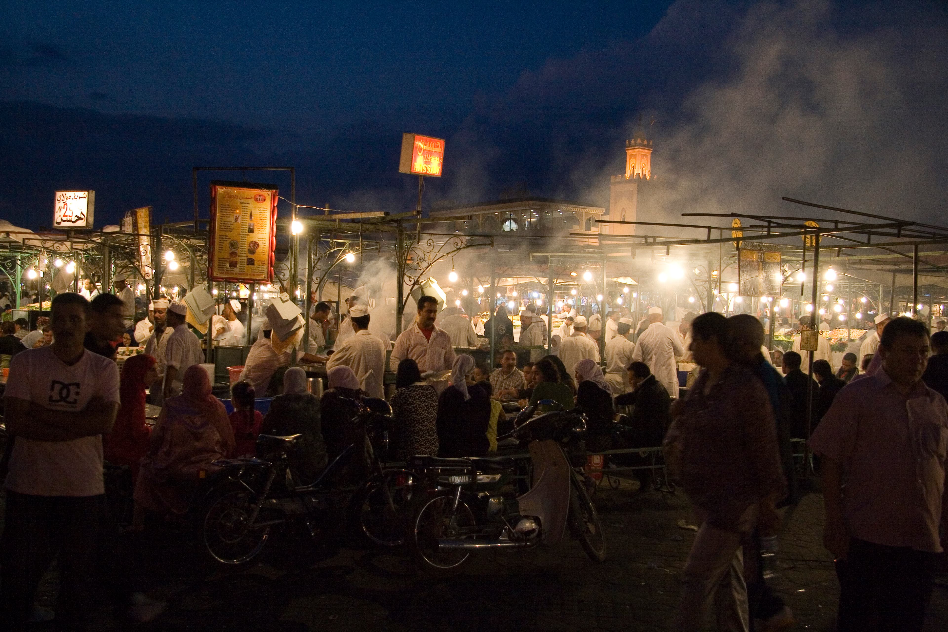 Jemaa el-Fna night market in Marrakech, steam rising from the food stalls and the Koutoubia minaret lit up in the background, close to the Marathon Village where marathon bib collection takes place