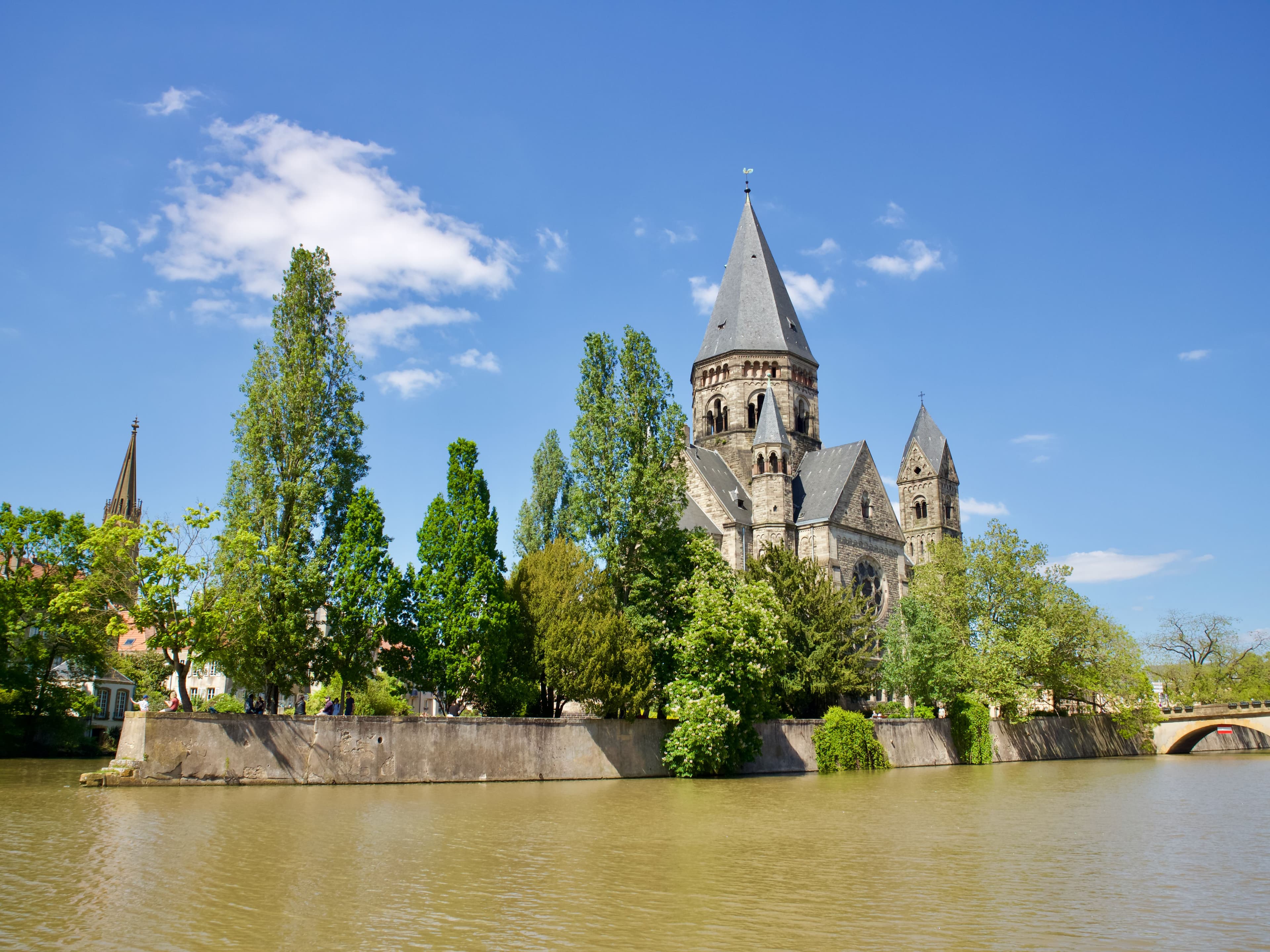 The Temple Neuf, a grey stone church with a tall slate spire, on its island in the Moselle in central Metz, behind a river wall and poplar trees; the Euro Marathon Metz passes it at km 5.7