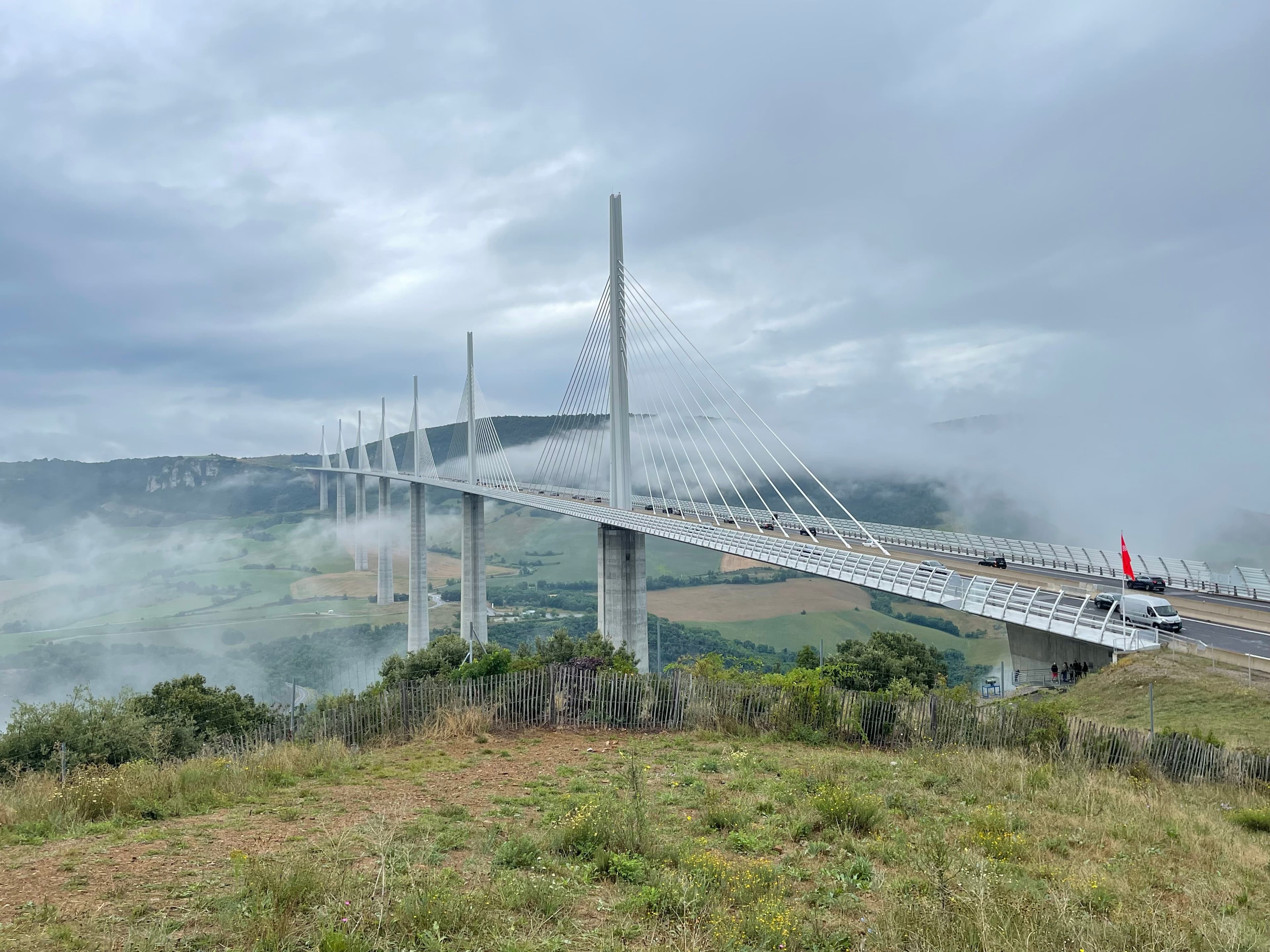 The Millau Viaduct in low cloud, its white pylons and cables crossing the Tarn valley south-west of Millau, seen from a grassy viewpoint; the marathon course runs the other way, north-east up the Tarn