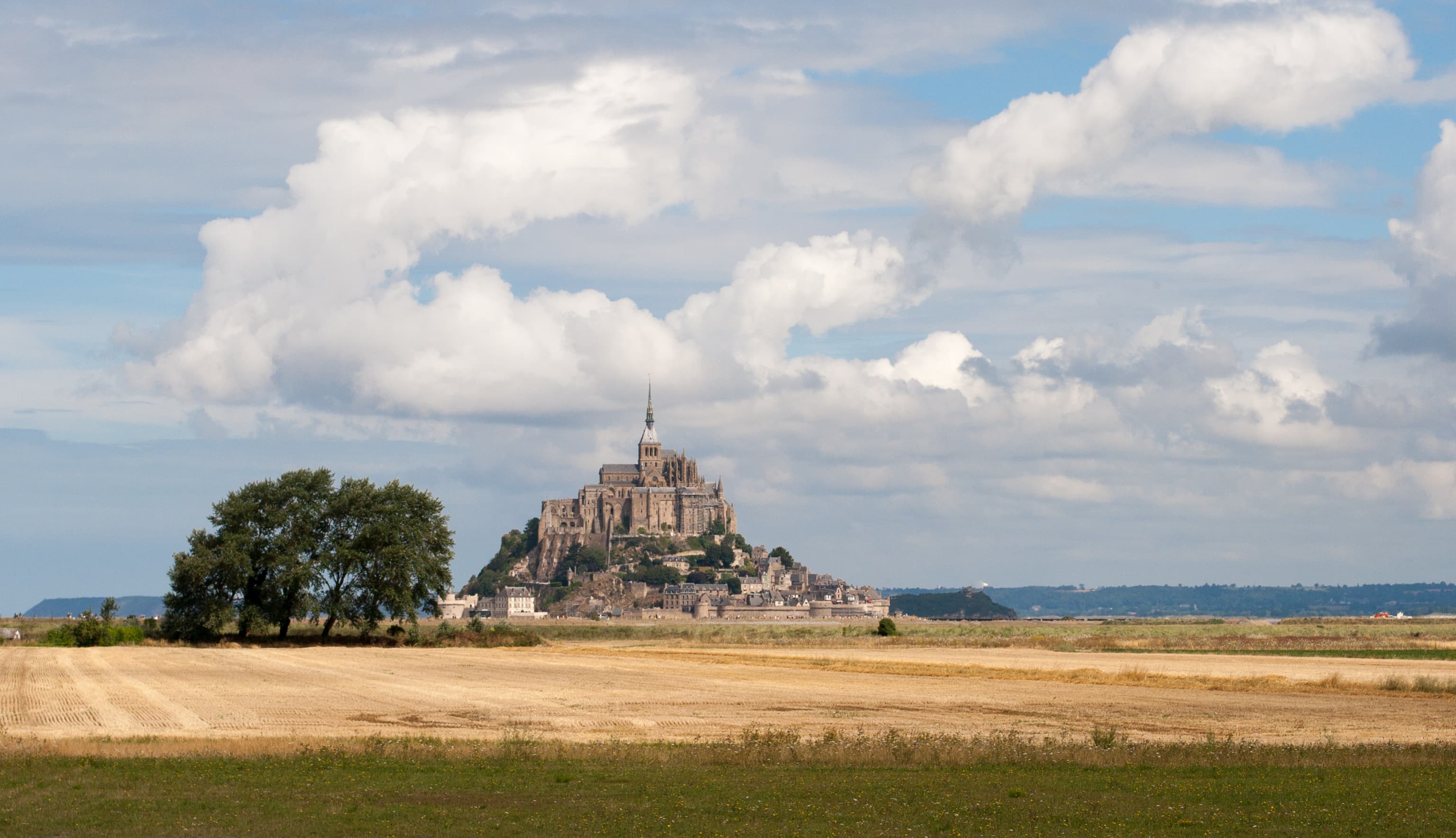 Mont Saint-Michel seen across harvested fields and salt meadows, the abbey spire rising above the walled village, with Tombelaine islet to the right and cumulus cloud over the bay.