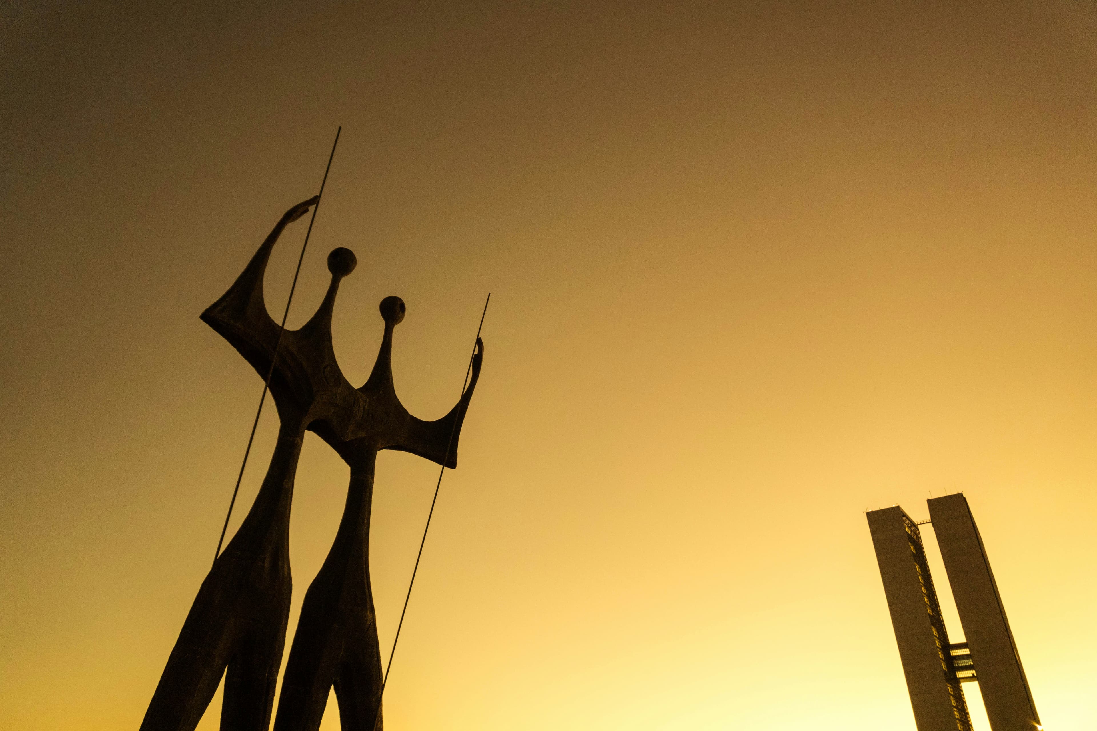 The Candangos sculpture in silhouette against a golden sky, with the twin towers of the National Congress on the right, in Brasília
