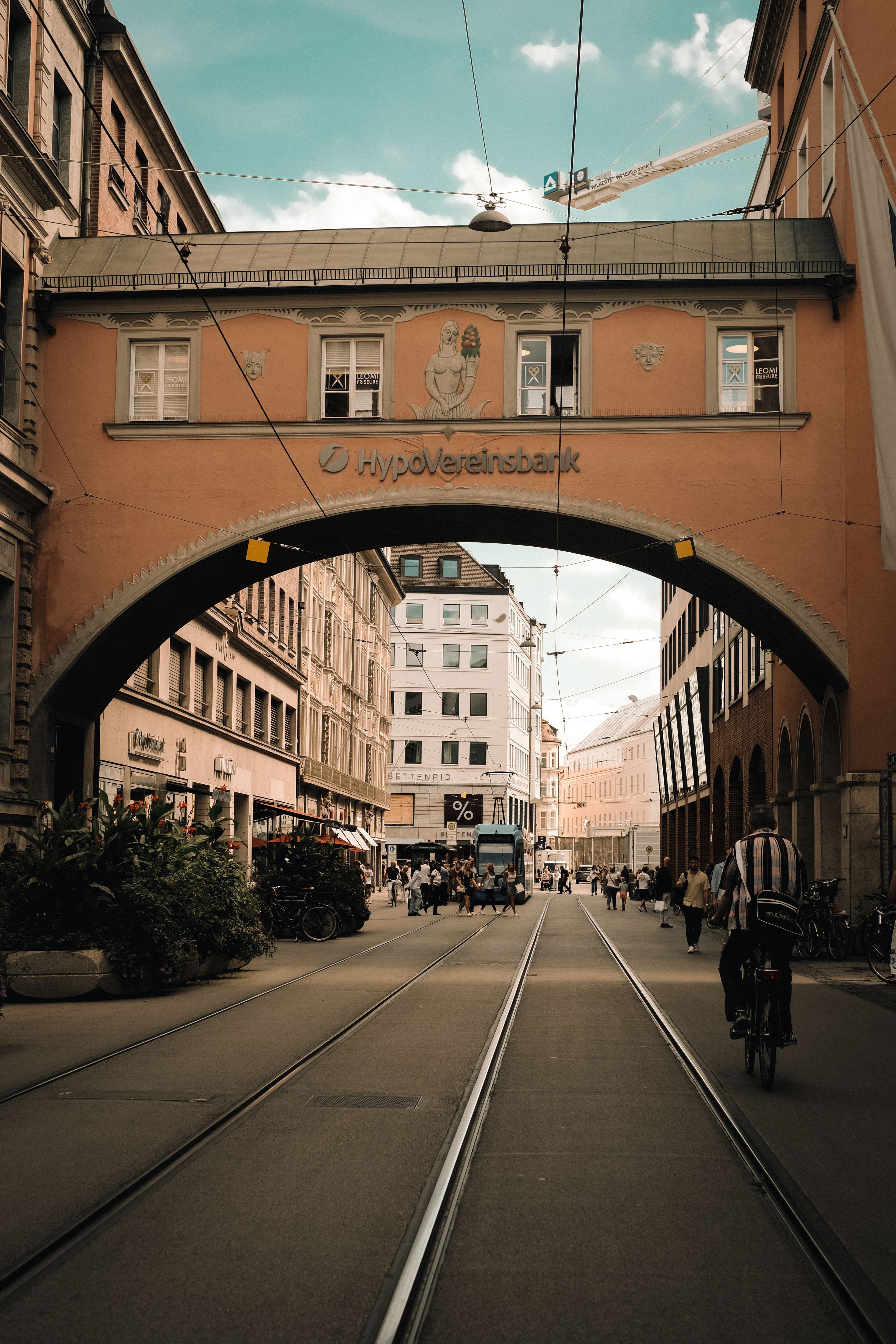 A tram street in central Munich running beneath an arched building bridge, with tracks curving away toward the old town. The marathon threads through these central streets around km 7 to 11.