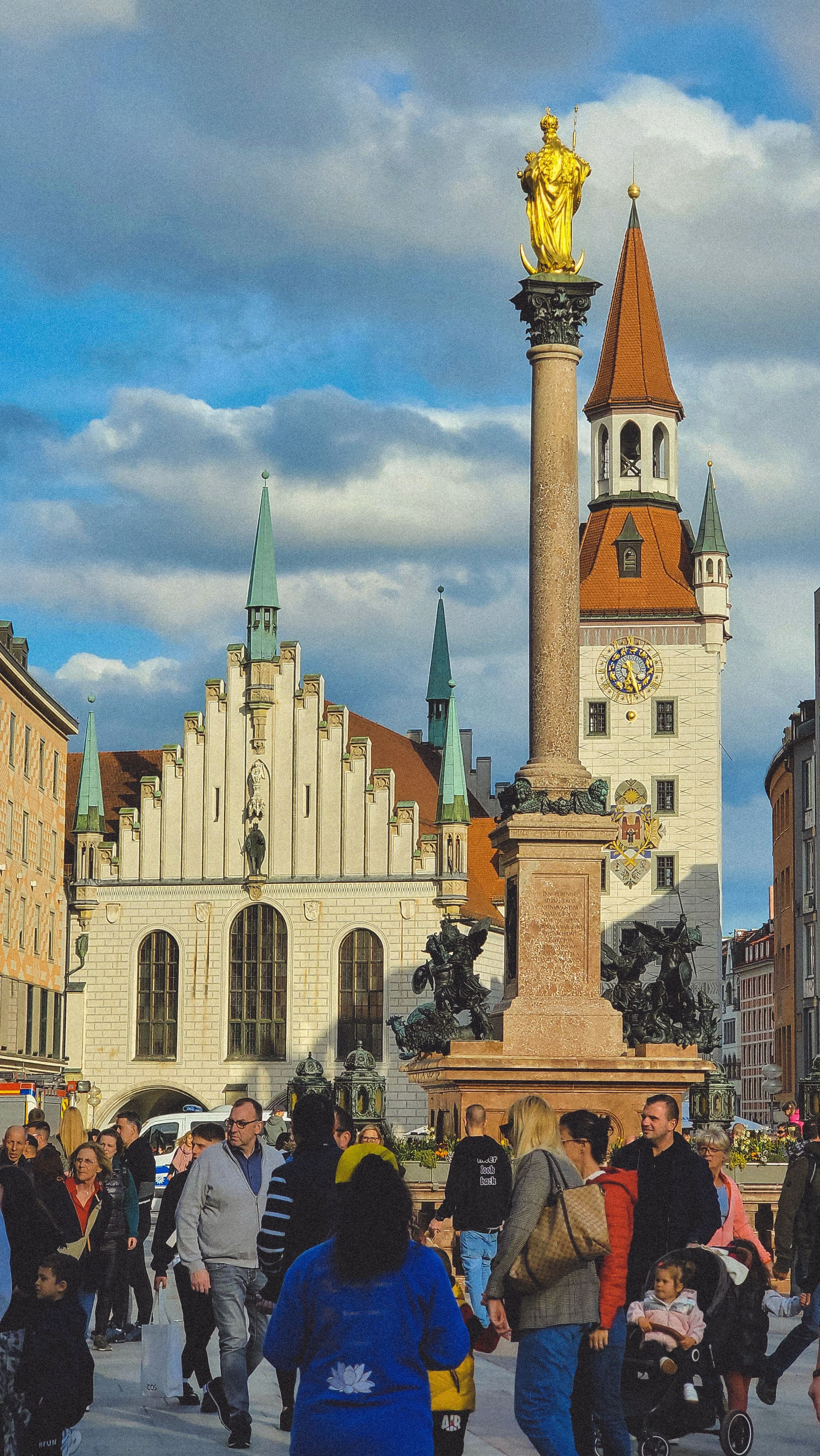 The Mariensäule column and the Altes Rathaus tower on Marienplatz, Munich, with crowds crossing the square. This is the densest spectator point on the marathon course.