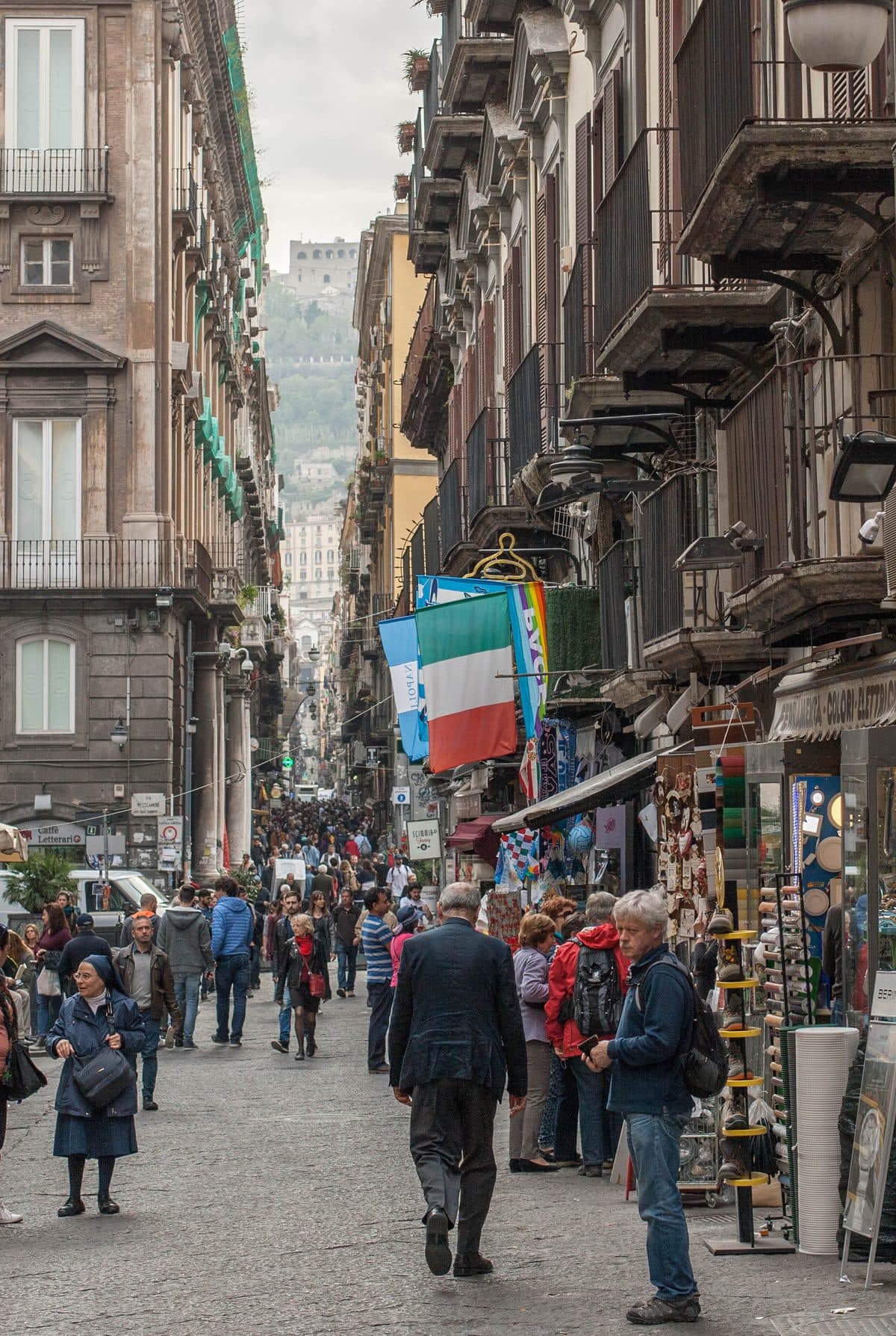 A crowded pedestrian street between tall palazzi in central Naples, with Italian and Napoli flags over the shops and a hilltop fortress at the far end