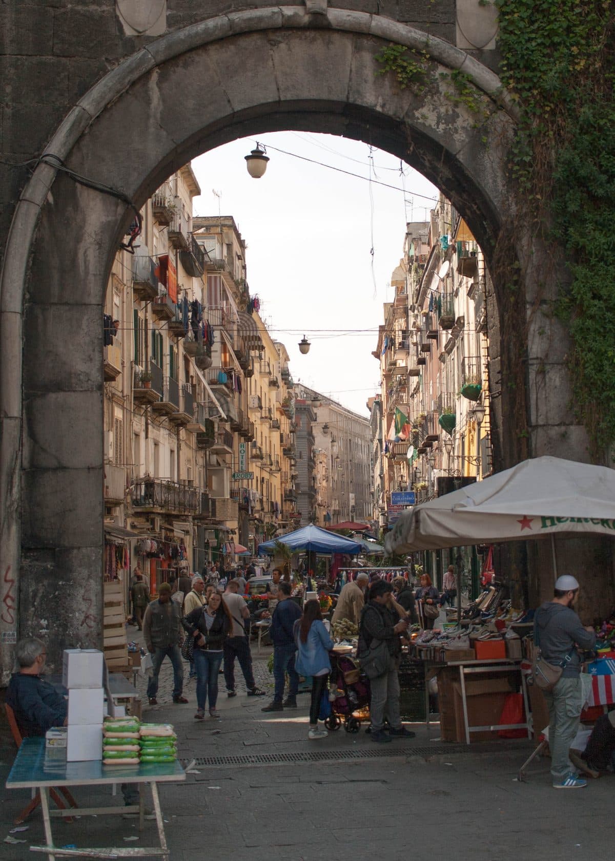 A weathered stone gateway framing a street market in Naples, with stalls, washing on balconies and shoppers beyond the arch