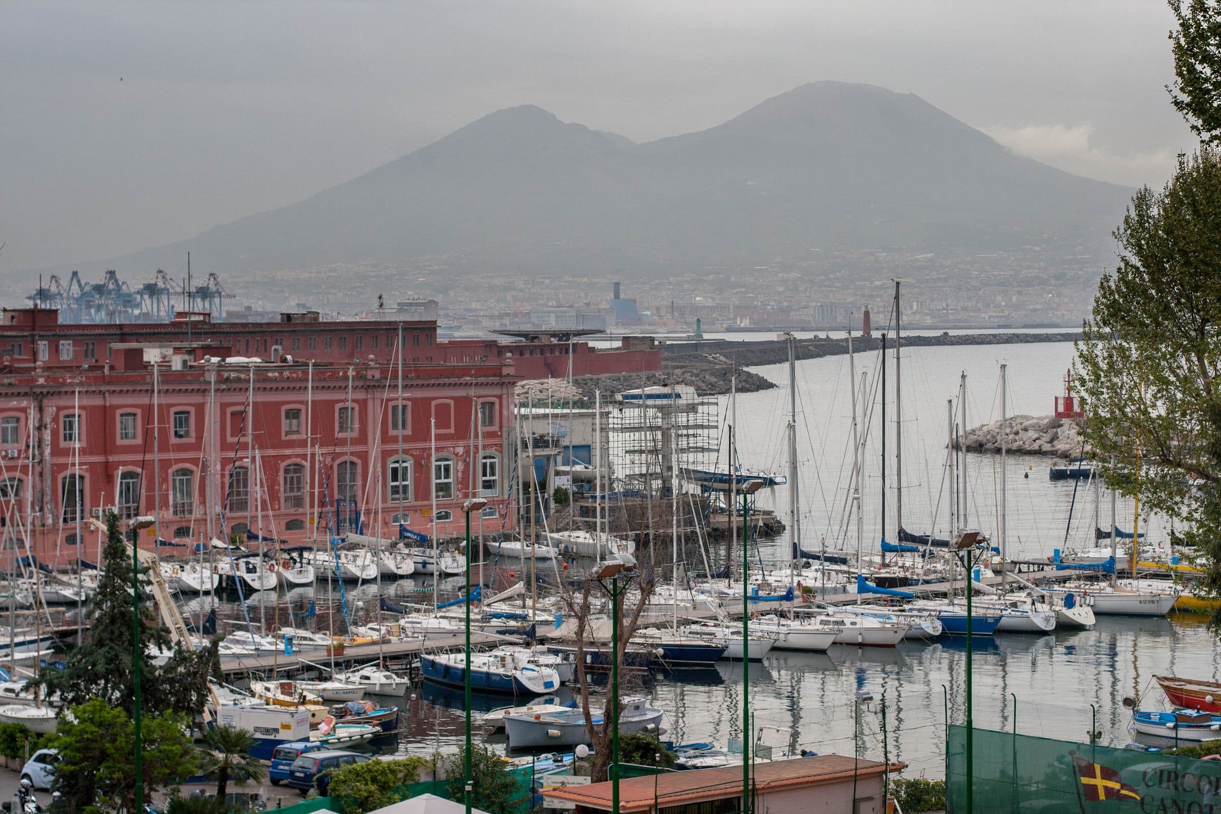 Moored yachts in a Naples marina with a red harbour-side building and Vesuvius rising through haze across the bay