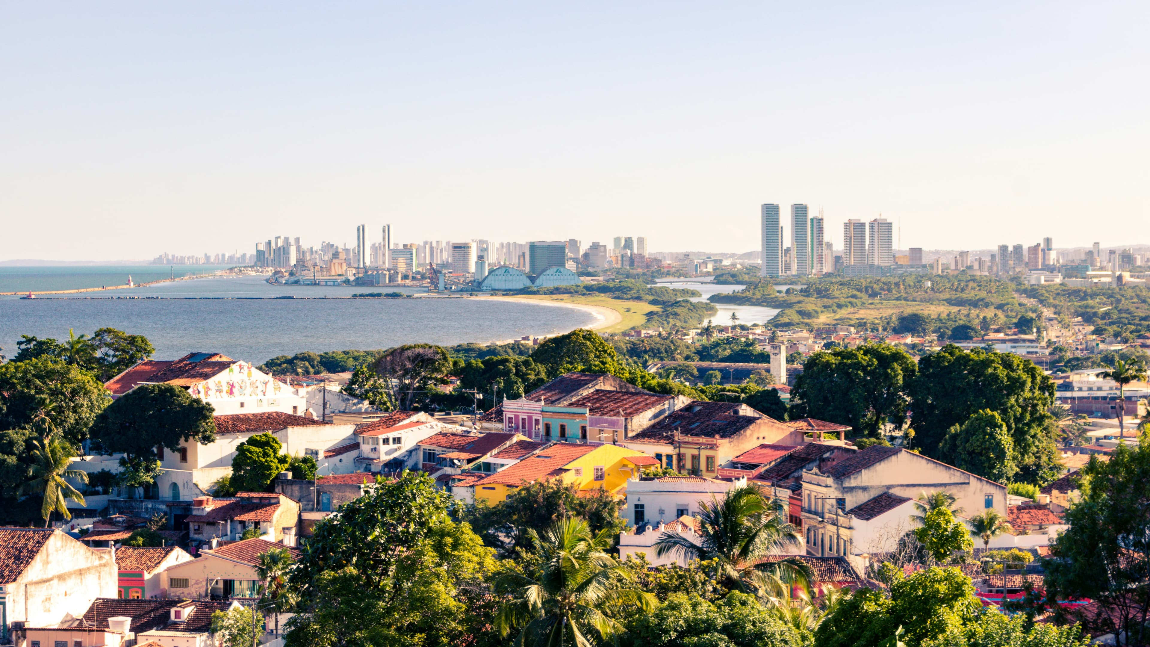 Terracotta roofs and colourful colonial houses of Olinda in the foreground, with Recife's harbour and high-rise skyline across the bay