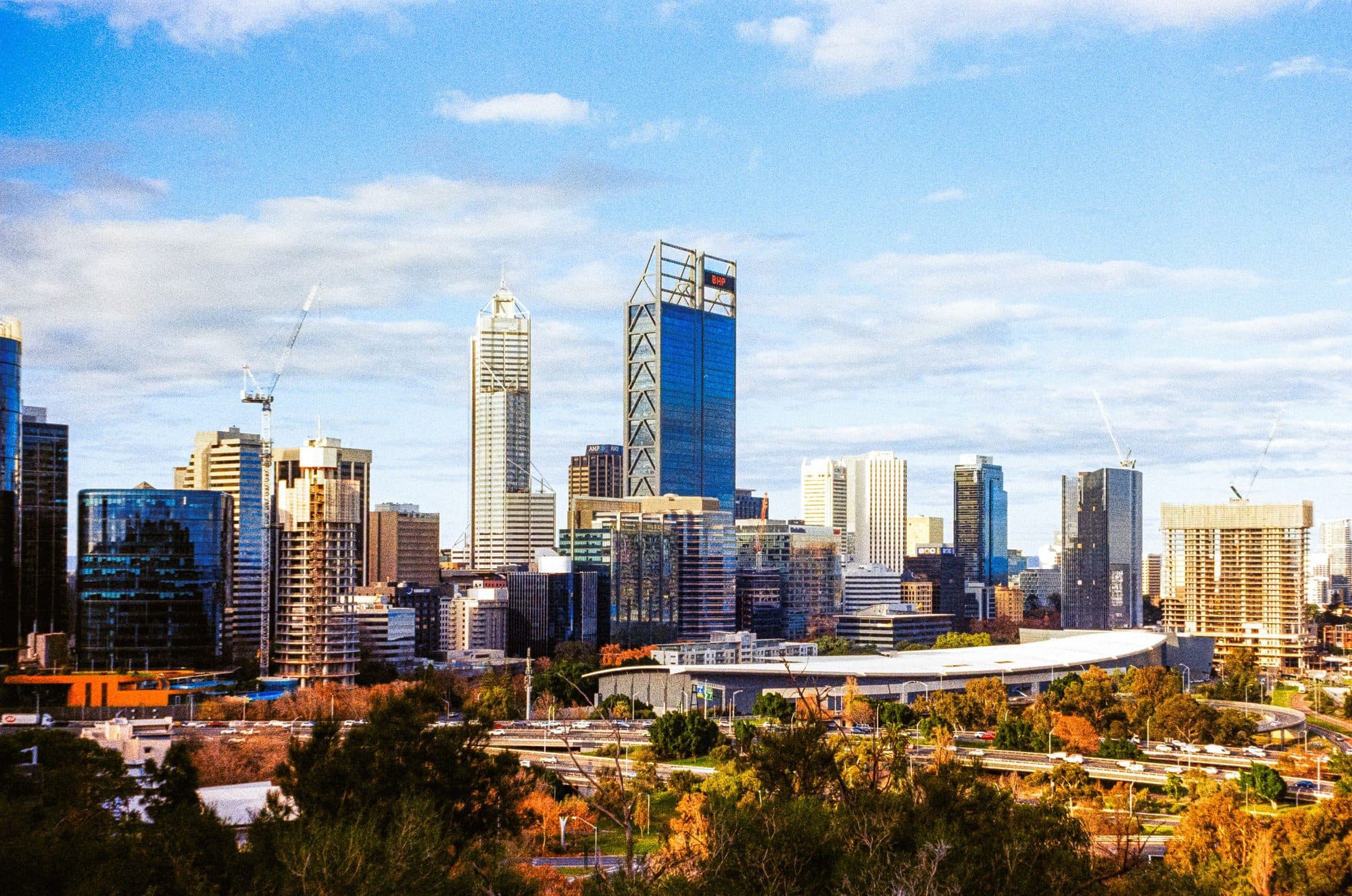 The Perth city skyline, including the BHP tower, seen across autumn-coloured trees and the freeway from Kings Park, beside the Mounts Bay Road end of the marathon route