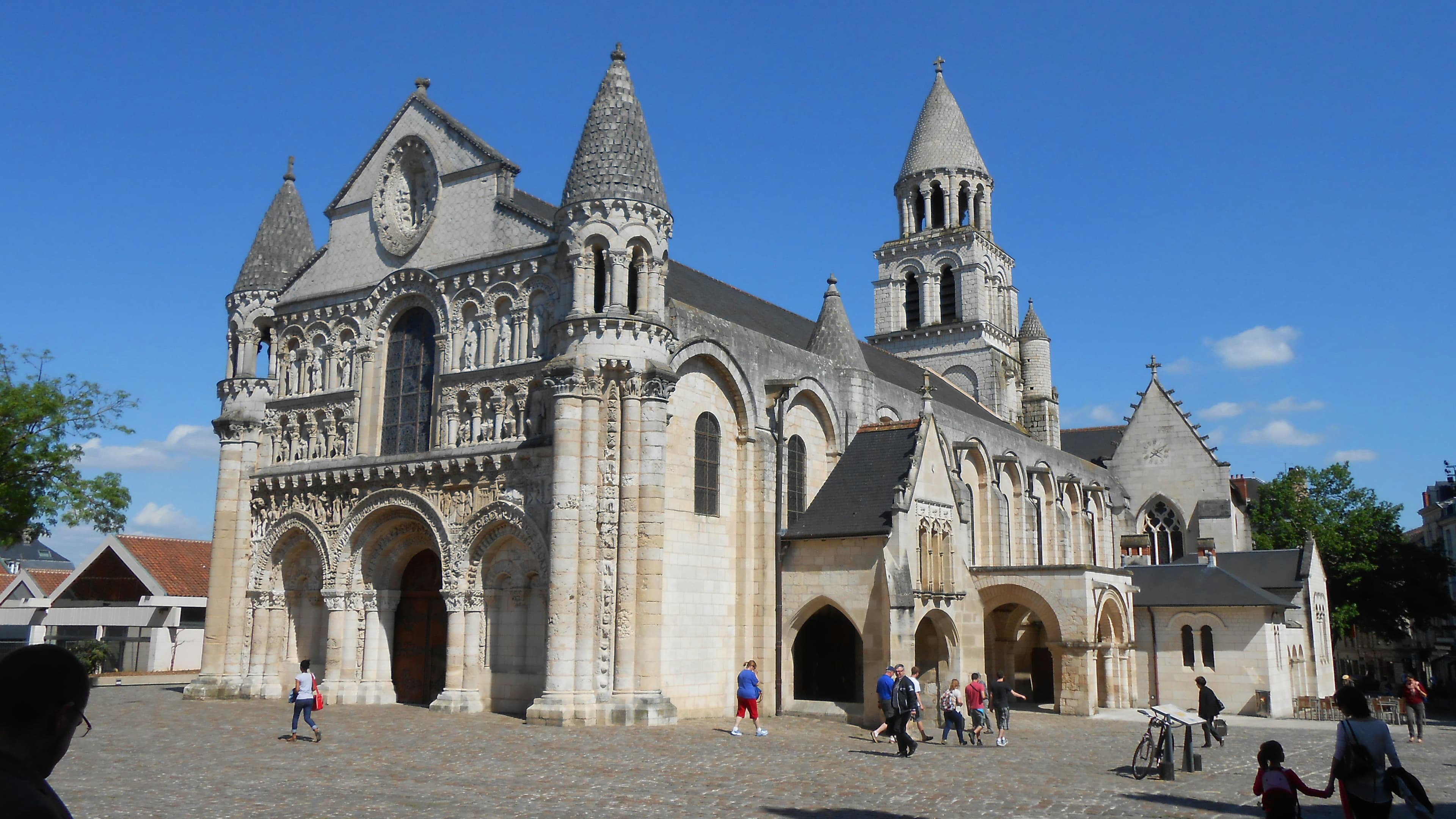 The carved Romanesque west front of Notre-Dame-la-Grande in Poitiers, with its two conical turrets and the bell tower behind, across a cobbled square under a blue sky; the marathon passes the church at km 1.2 and again at km 14.8