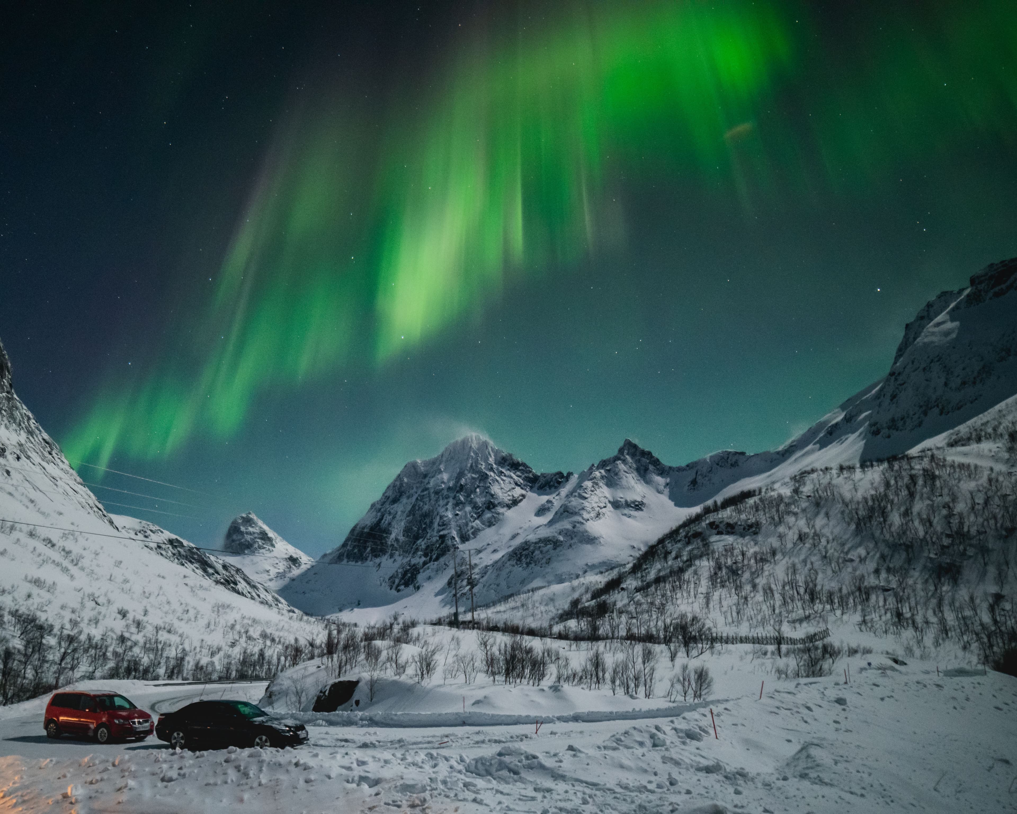 Vivid green northern lights sweeping across the night sky above snow-covered mountains near Tromsø, with two cars parked in a snow-filled valley below