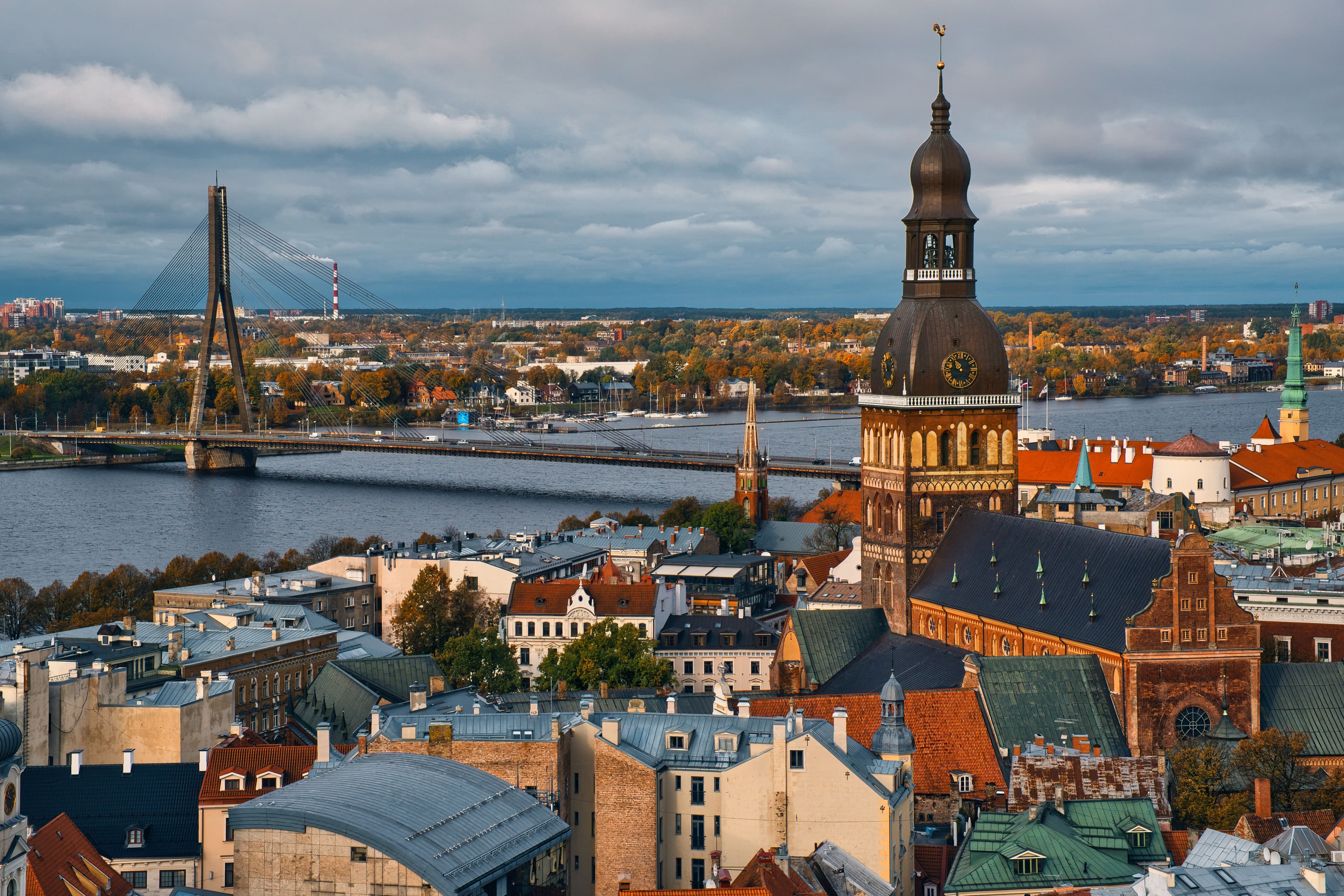 Aerial view of Riga's Old Town showing the Dome Cathedral with its copper dome, red-tiled rooftops, the Daugava River, and Vanšu Bridge in autumn light