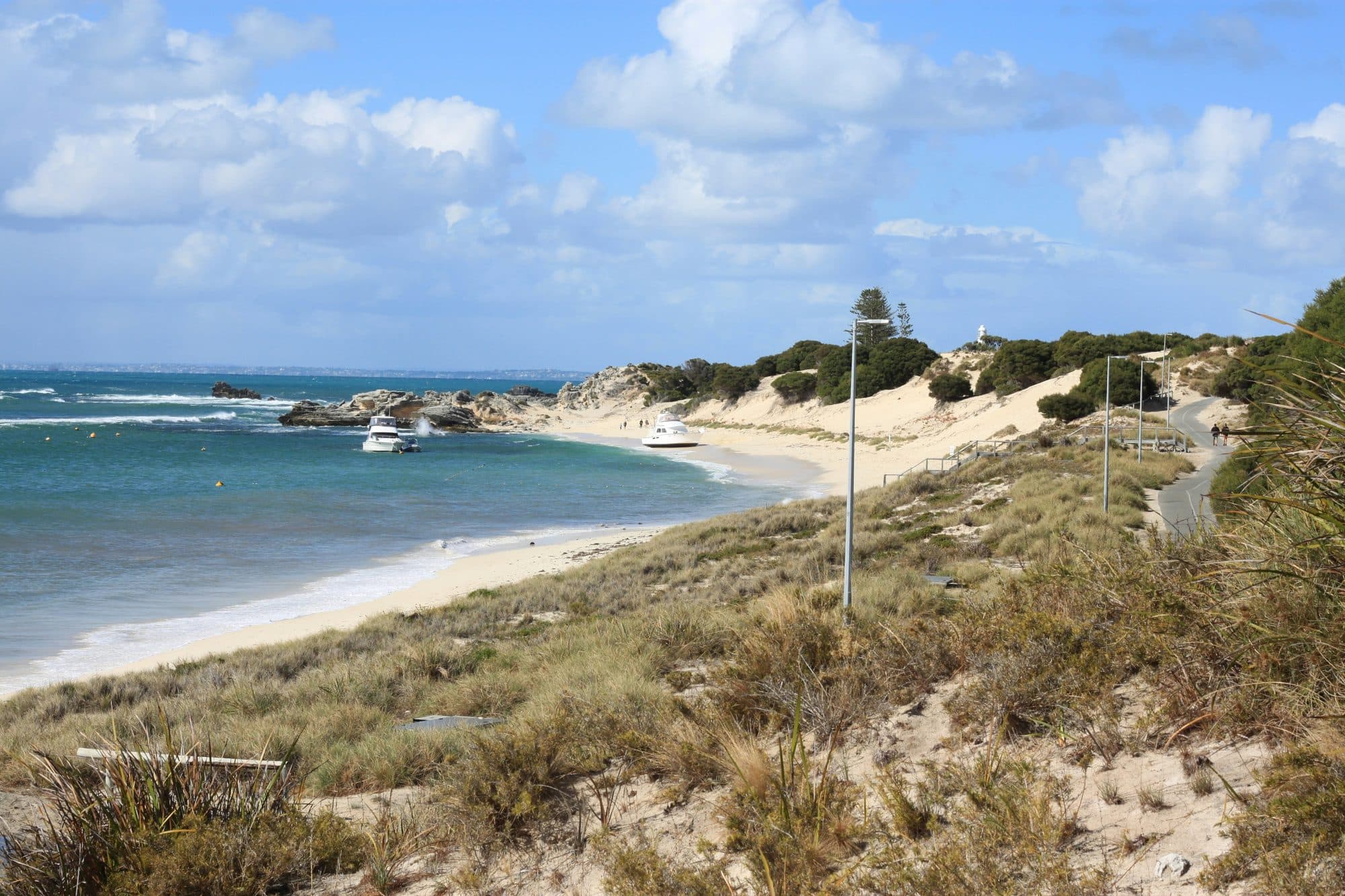 A sandy bay with turquoise water, moored boats, dune grass, a paved coastal path and a distant white lighthouse on Rottnest Island