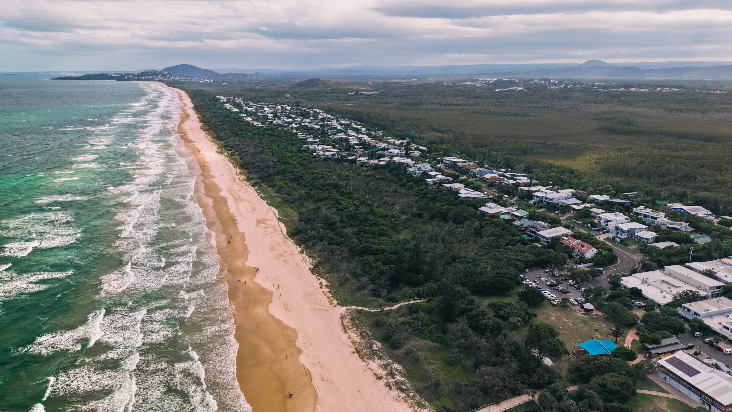 Aerial view of a long surf beach on the northern Sunshine Coast, with low dunes, beachside houses behind the banksia scrub and headlands on the horizon