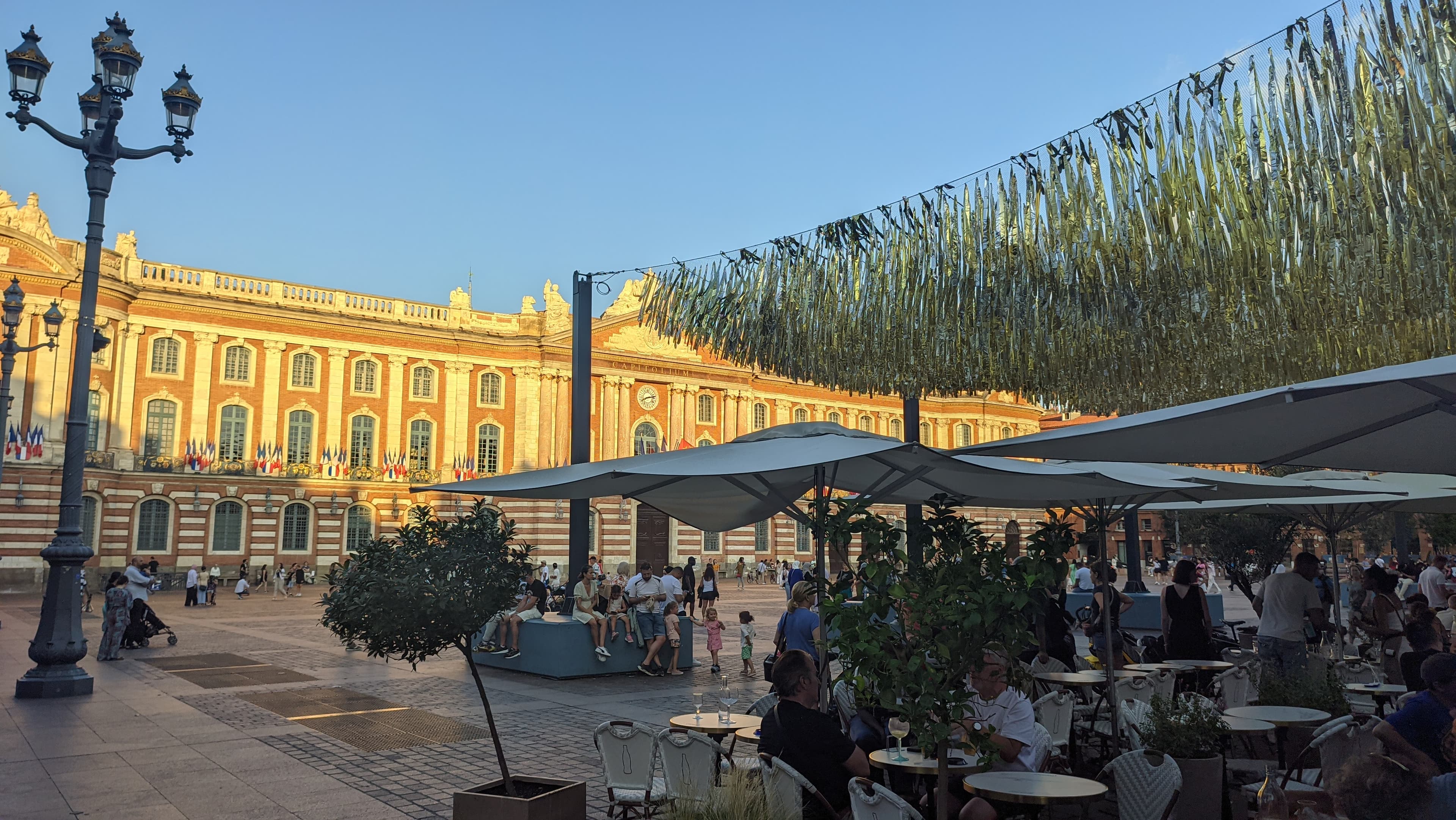 Evening light on the pink-brick facade of the Capitole in Toulouse, French flags along its balcony and cafe terraces on the square below, the site of the Marathon des Geants race village and the course at about km 3