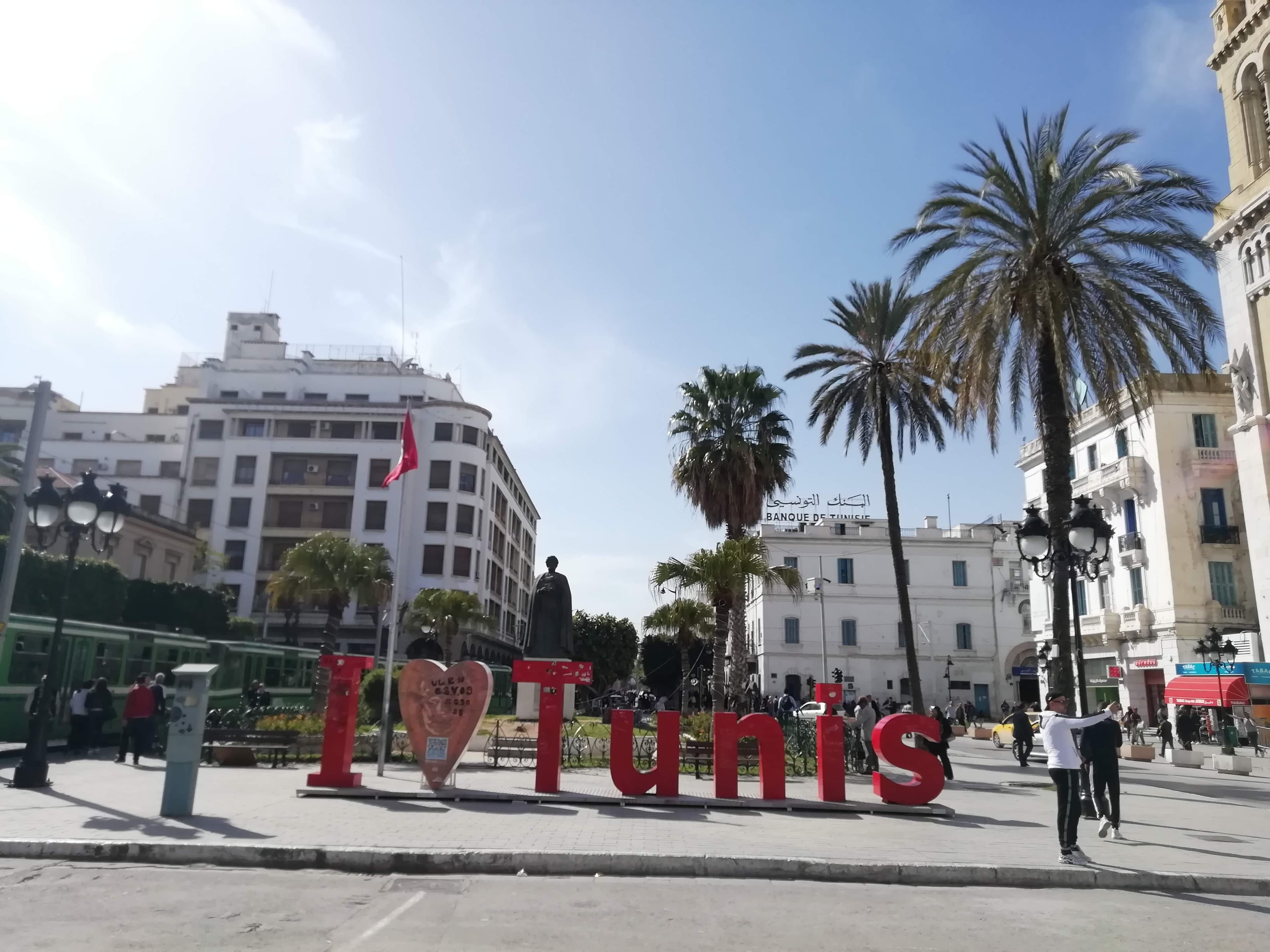 The red I Love Tunis sign on the square beside the Ibn Khaldun statue on Avenue Habib Bourguiba, central Tunis, with palm trees and a green TGM-style tram behind it. The marathon finishes on this avenue.