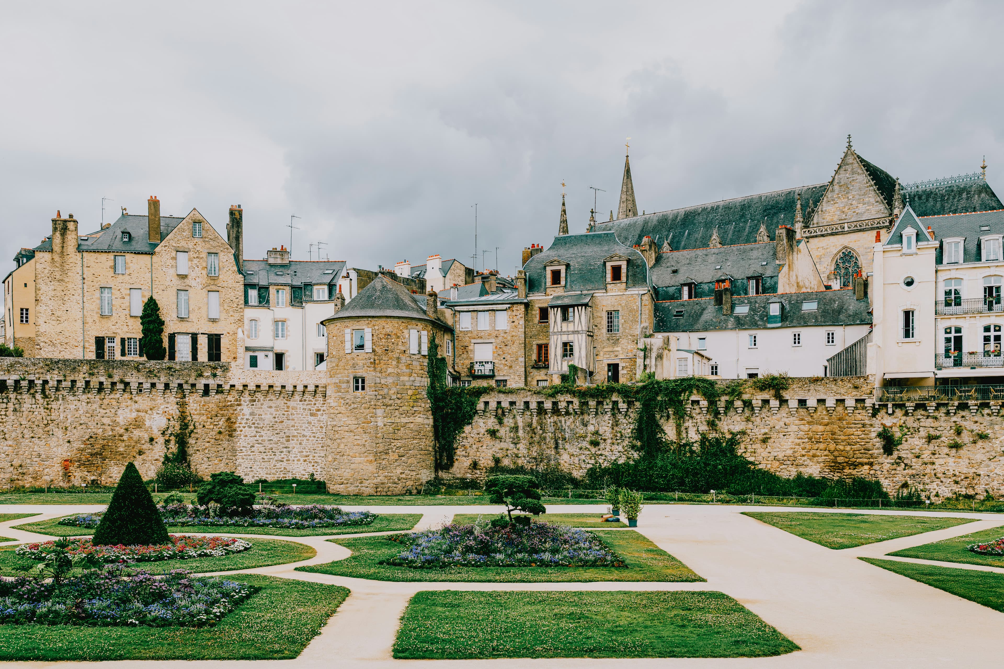 The medieval ramparts of Vannes with a round stone tower and the old town and cathedral roofs rising behind them, seen across the formal flower beds of the Jardins des Remparts, the setting for the Marathon de Vannes start on Rue Francis Decker