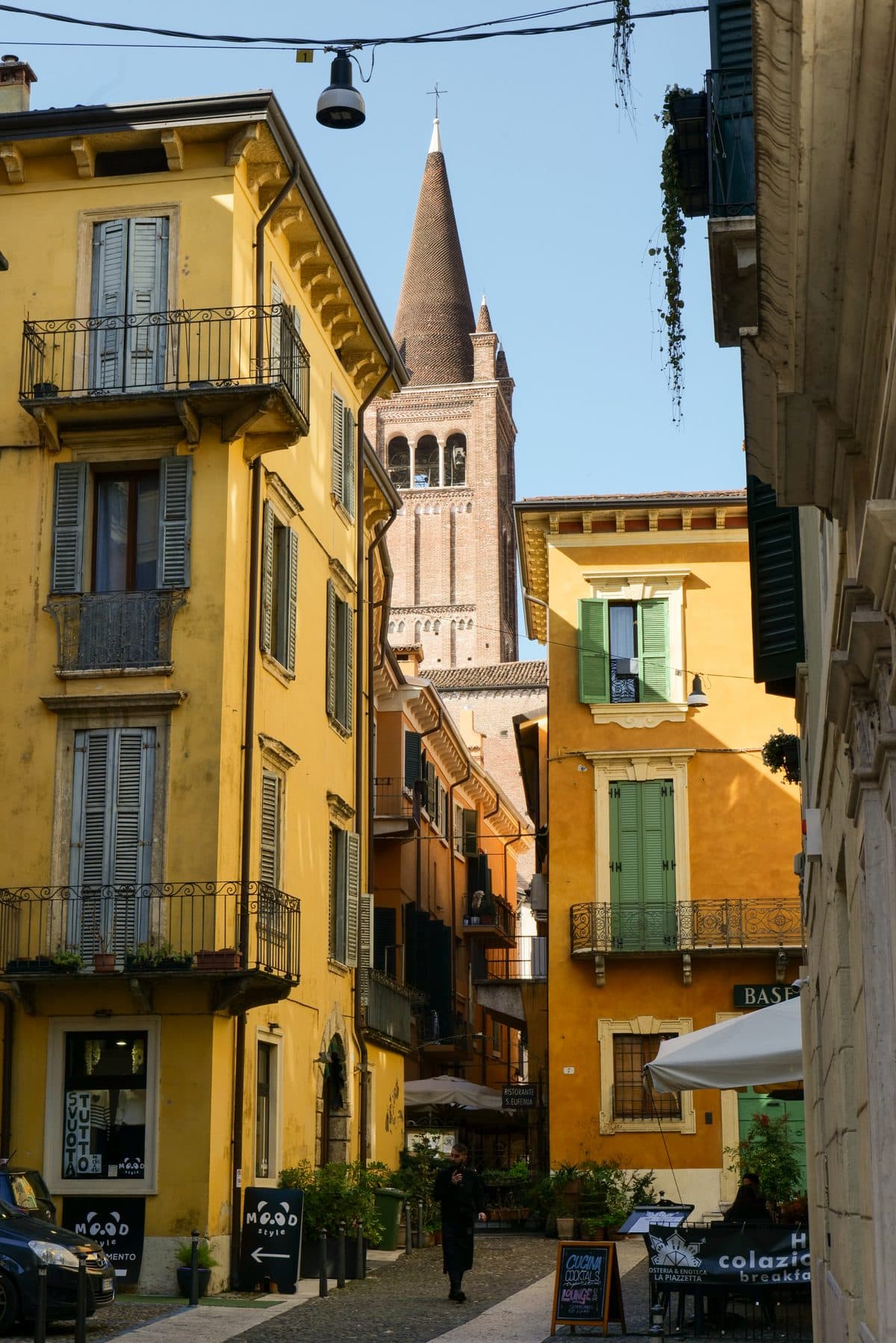 Ochre and yellow houses with green shutters on a narrow Verona lane, with a brick bell tower and conical spire rising behind them
